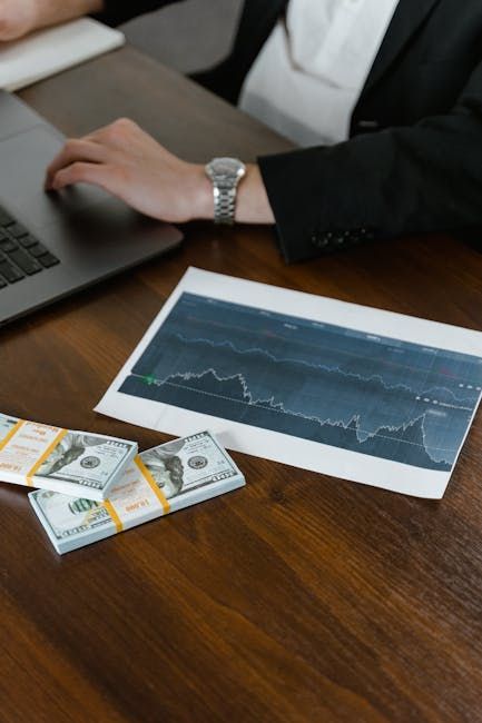 Person in a suit at a desk with a laptop, stacks of cash, and a graph showing a downward trend.