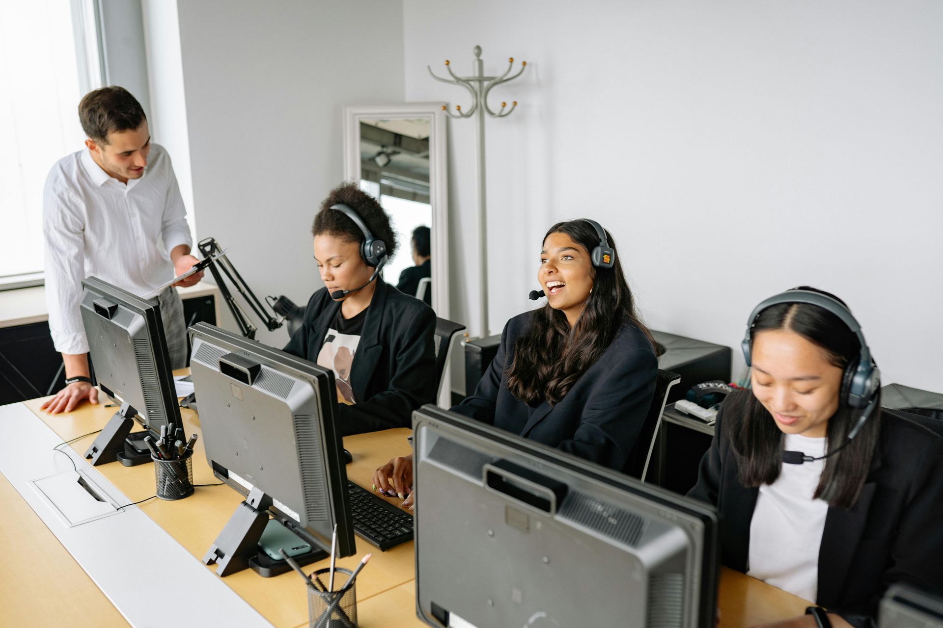 A supervisor stands beside three employees wearing headsets at their desks in an office, working on computers.