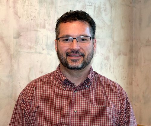 A man wearing glasses and a yellow shirt is smiling for the camera