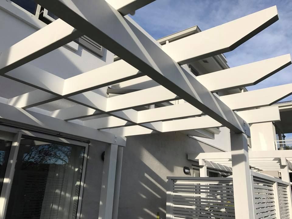 White pergola over an outdoor patio, casting shadows on the light gray building and fence. White pergola over an outdoor patio, casting shadows on the light gray building and fence.