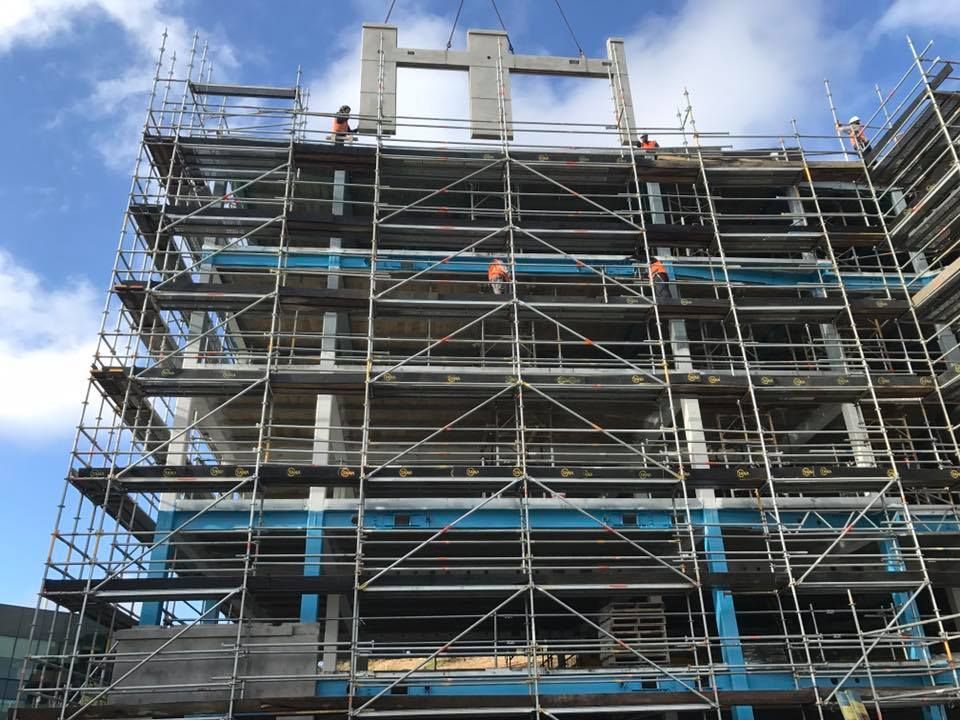 Construction workers on scaffolding, lifting a large concrete beam onto a multi-story building frame, under a blue sky. Construction workers on scaffolding, lifting a large concrete beam onto a multi-story building frame, under a blue sky.