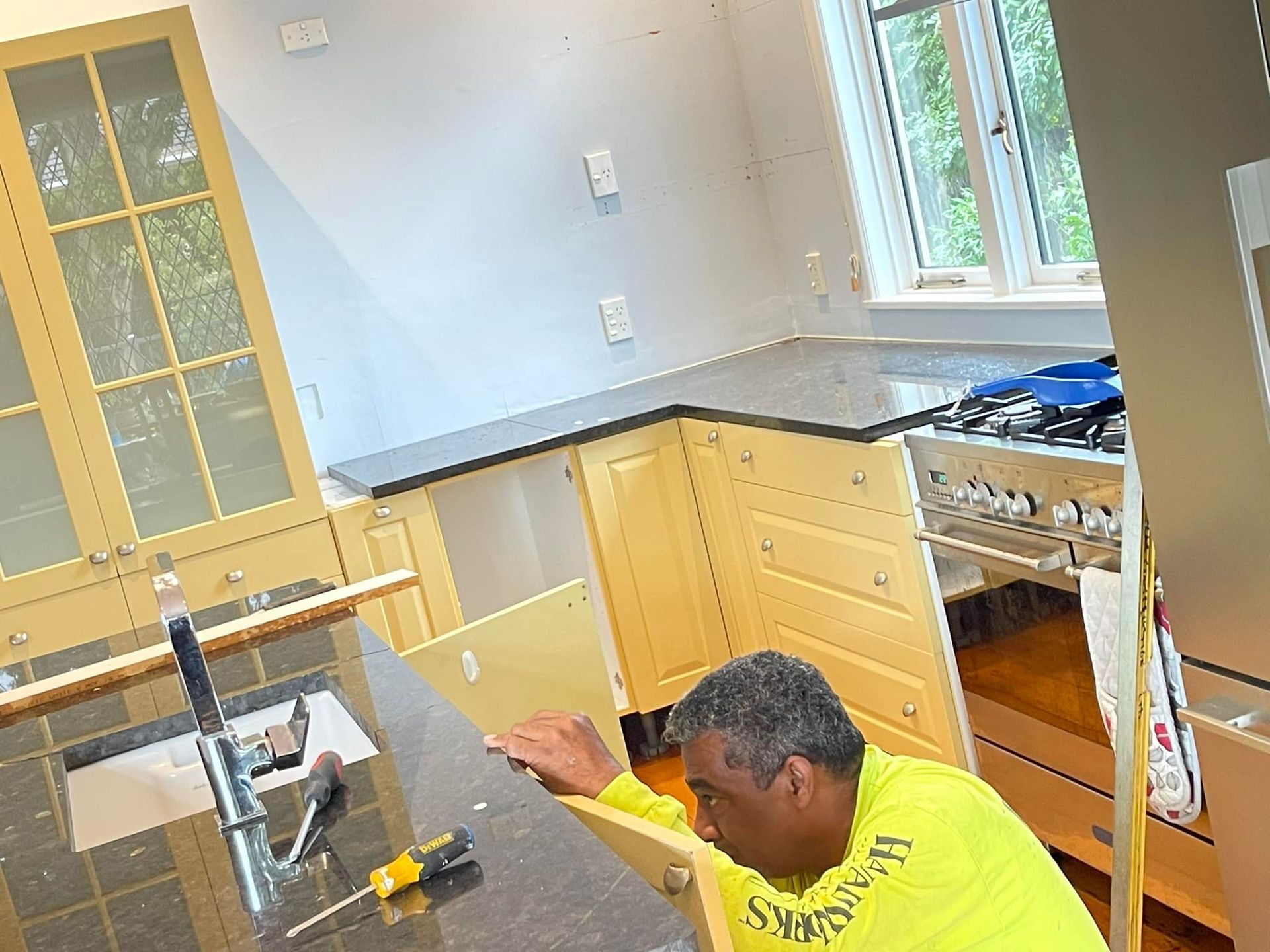 A person installing a cabinet in a kitchen, wearing a yellow shirt, with black countertops and a window. A person installing a cabinet in a kitchen, wearing a yellow shirt, with black countertops and a window.