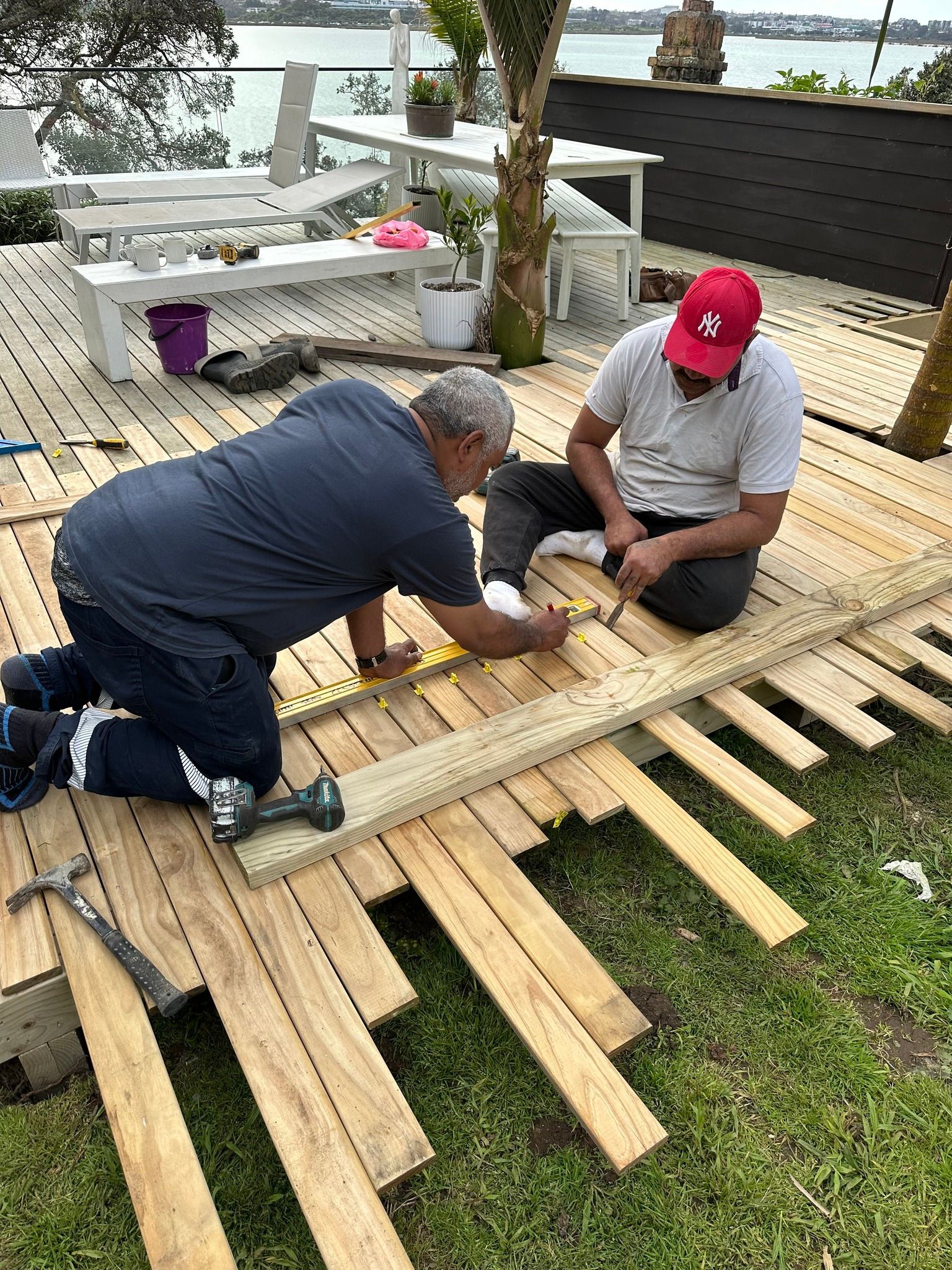 Two men building a wooden deck outdoors. One kneels and works with a tool. The other sits, also working.