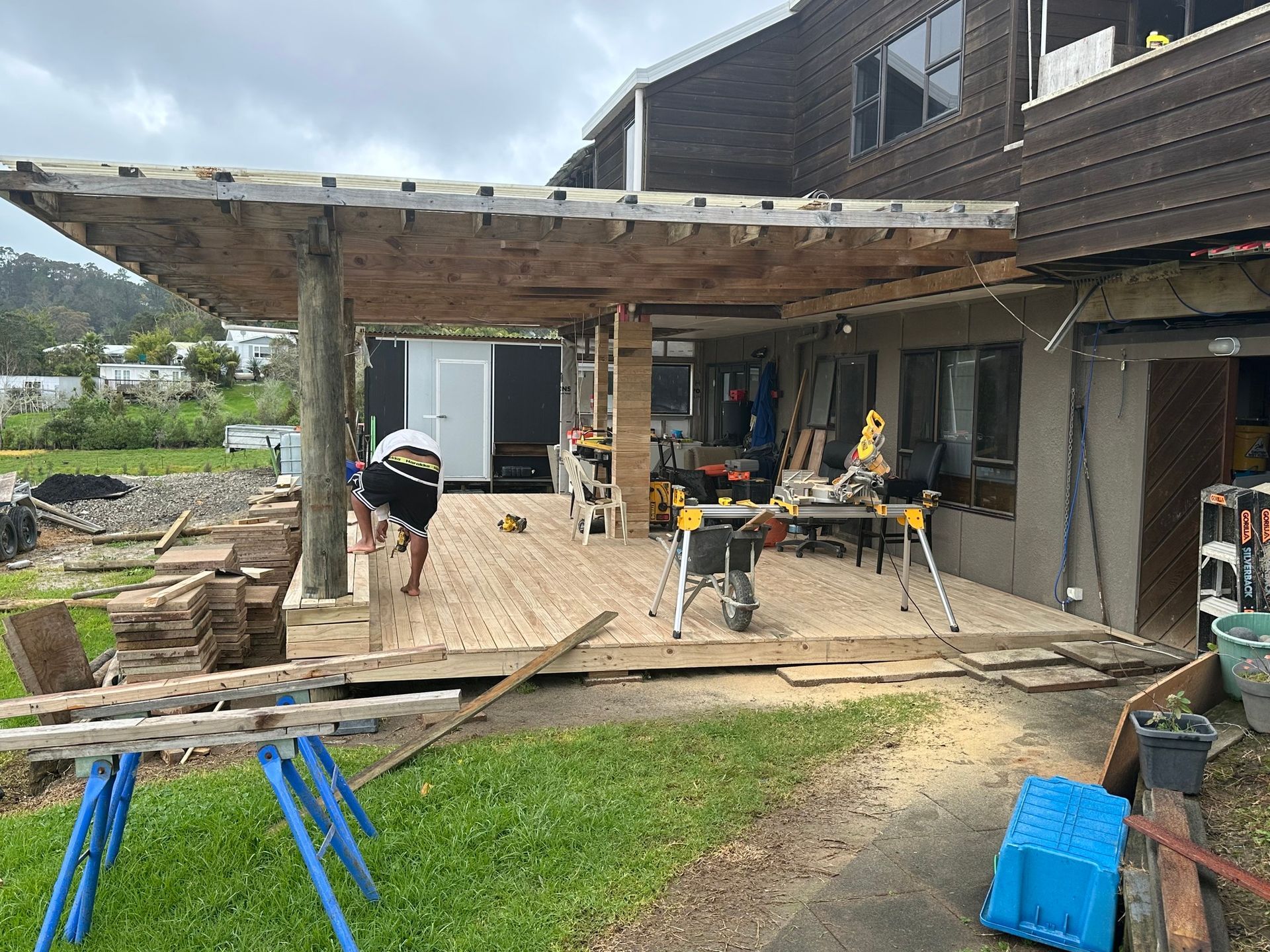Deck construction in progress; person working on wooden deck with tools. Brown wood and green grass.