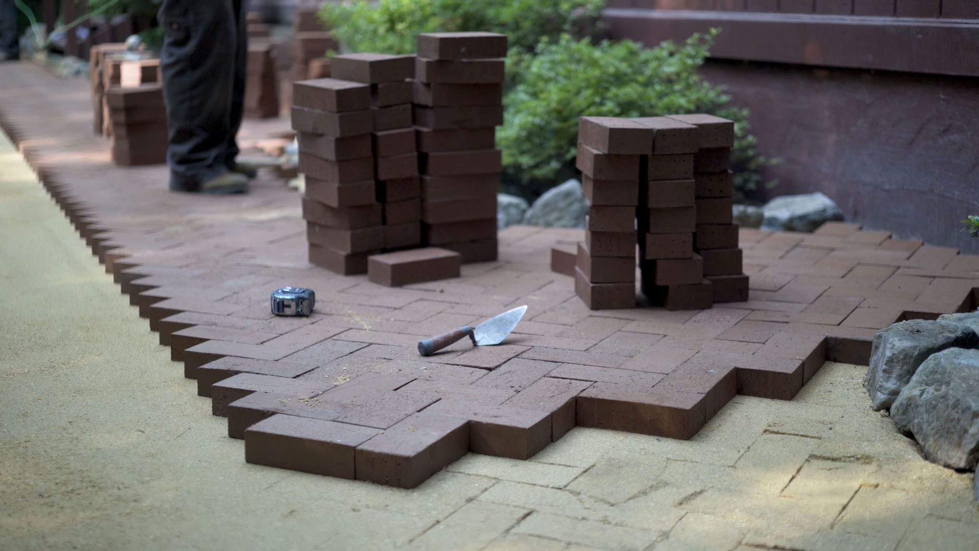 Person laying dark bricks for a walkway. Several stacks of bricks and tools are present.