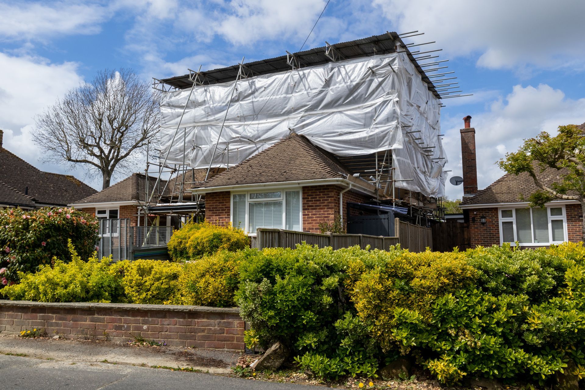 House covered in scaffolding and white sheeting for renovation, surrounded by bushes and other houses.