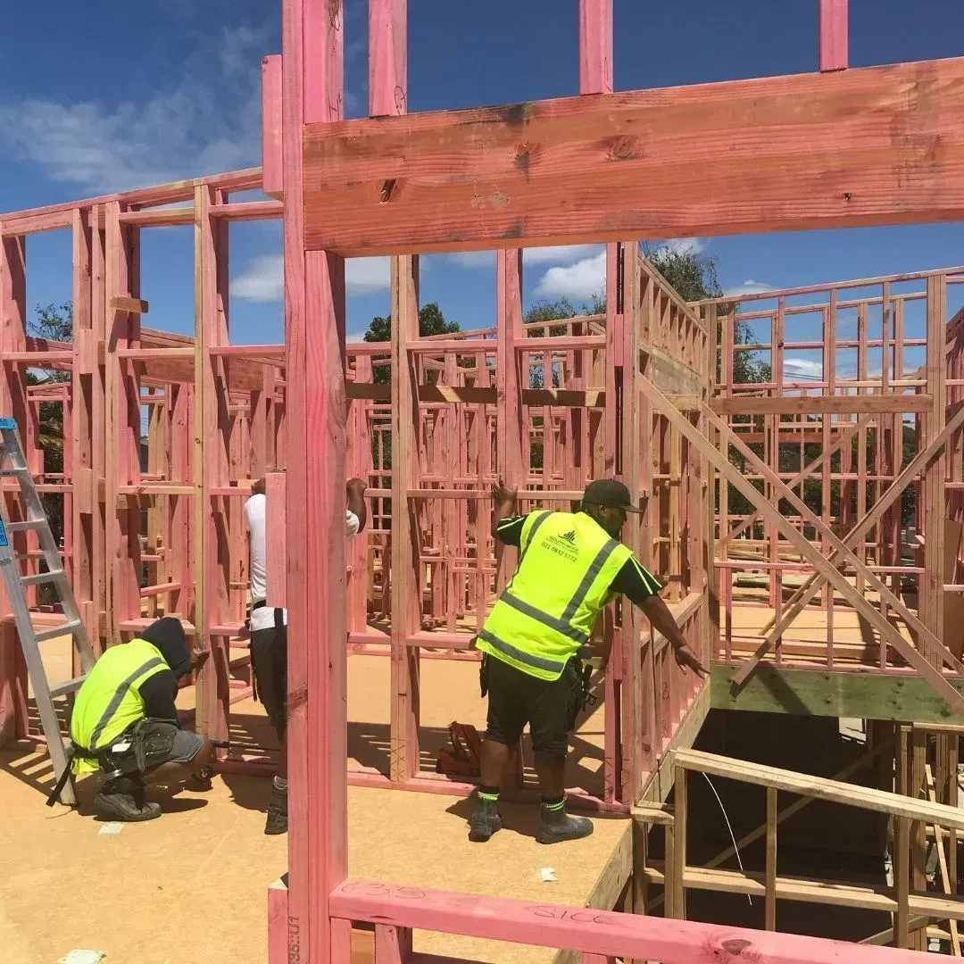 Construction workers framing a building. Wooden structure with pink treated lumber. Two workers in safety vests. Sunny day.