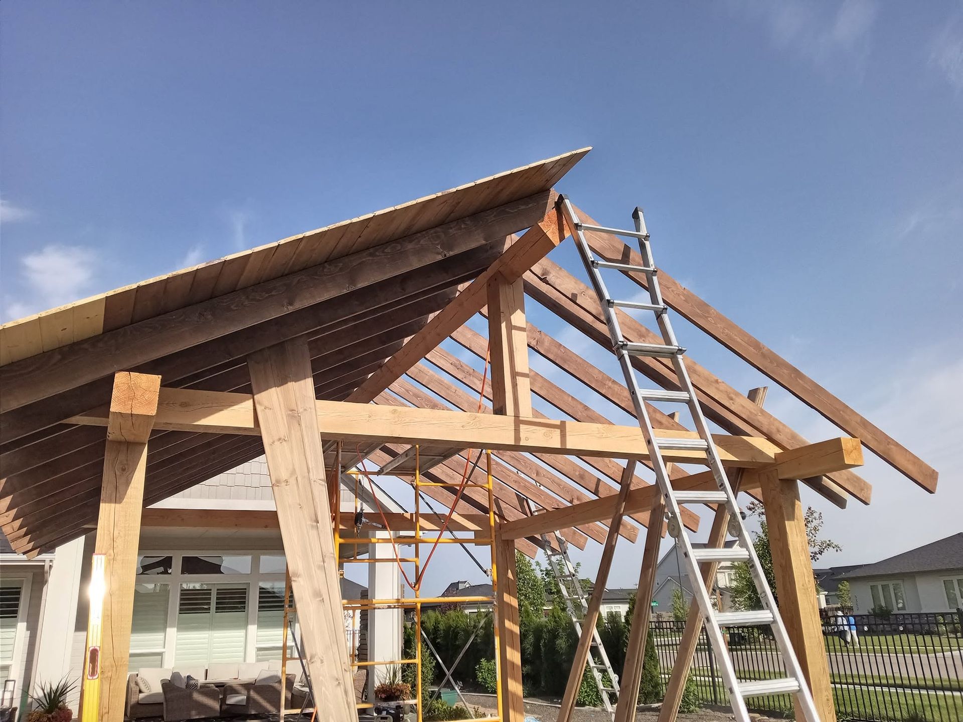Wooden pavilion frame under construction with a ladder and exposed roof beams against a blue sky