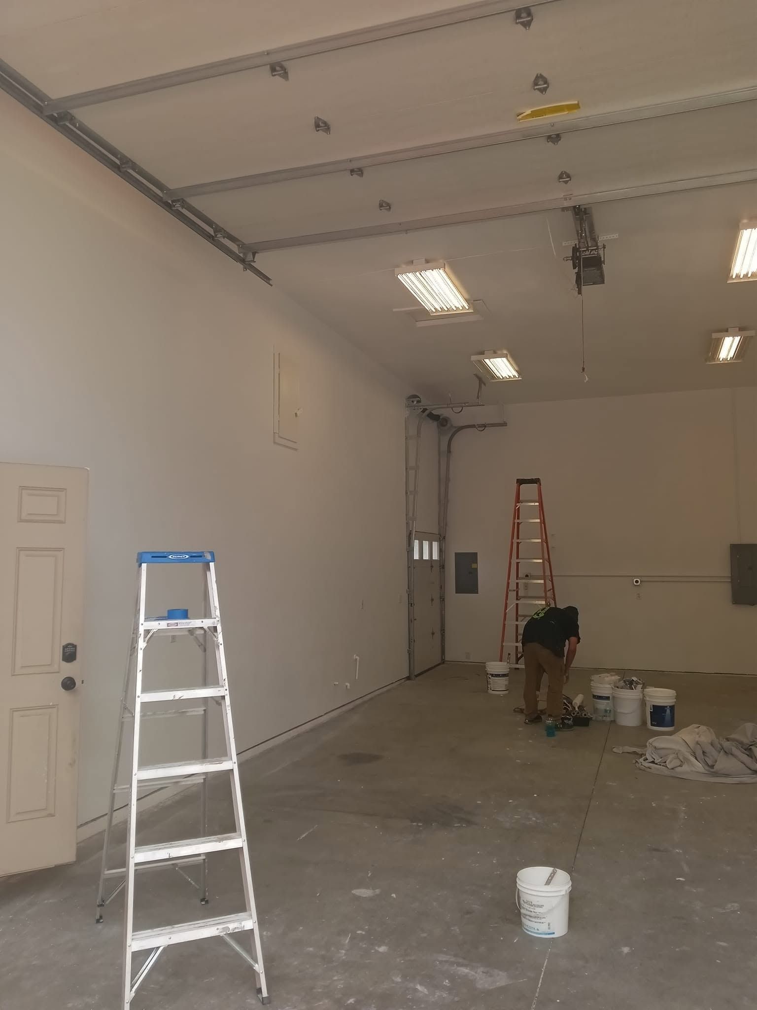 Interior of a garage or workshop under renovation, with ladders, tools, and a person working near the back wall.