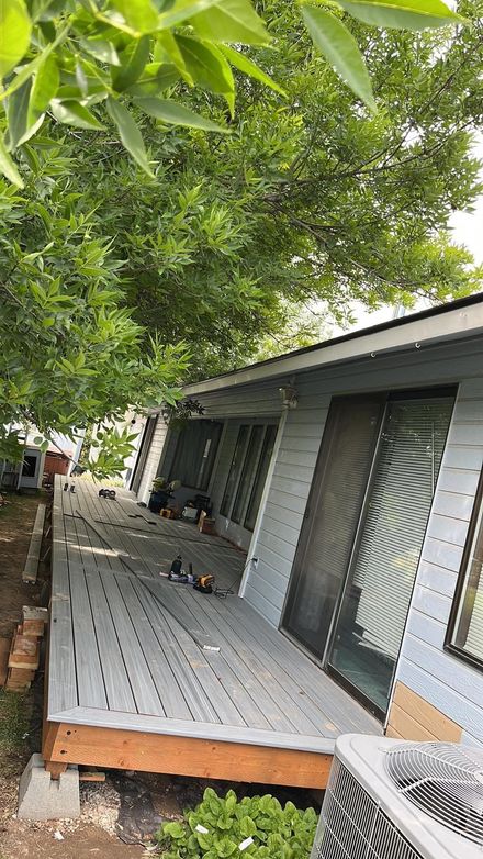 Gray house with a wooden porch under leafy green trees, viewed at an angle.