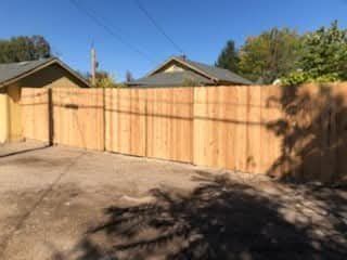 Wooden privacy fence enclosing a sunlit driveway beside a house, with tree shadows in the foreground.