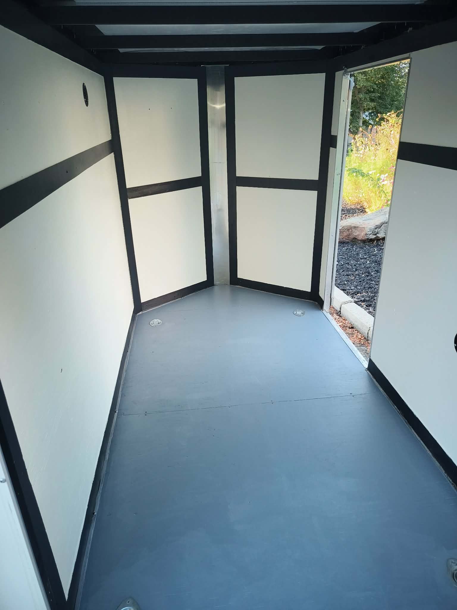 Empty narrow trailer interior with white walls, black trim, and an open door to the outside.