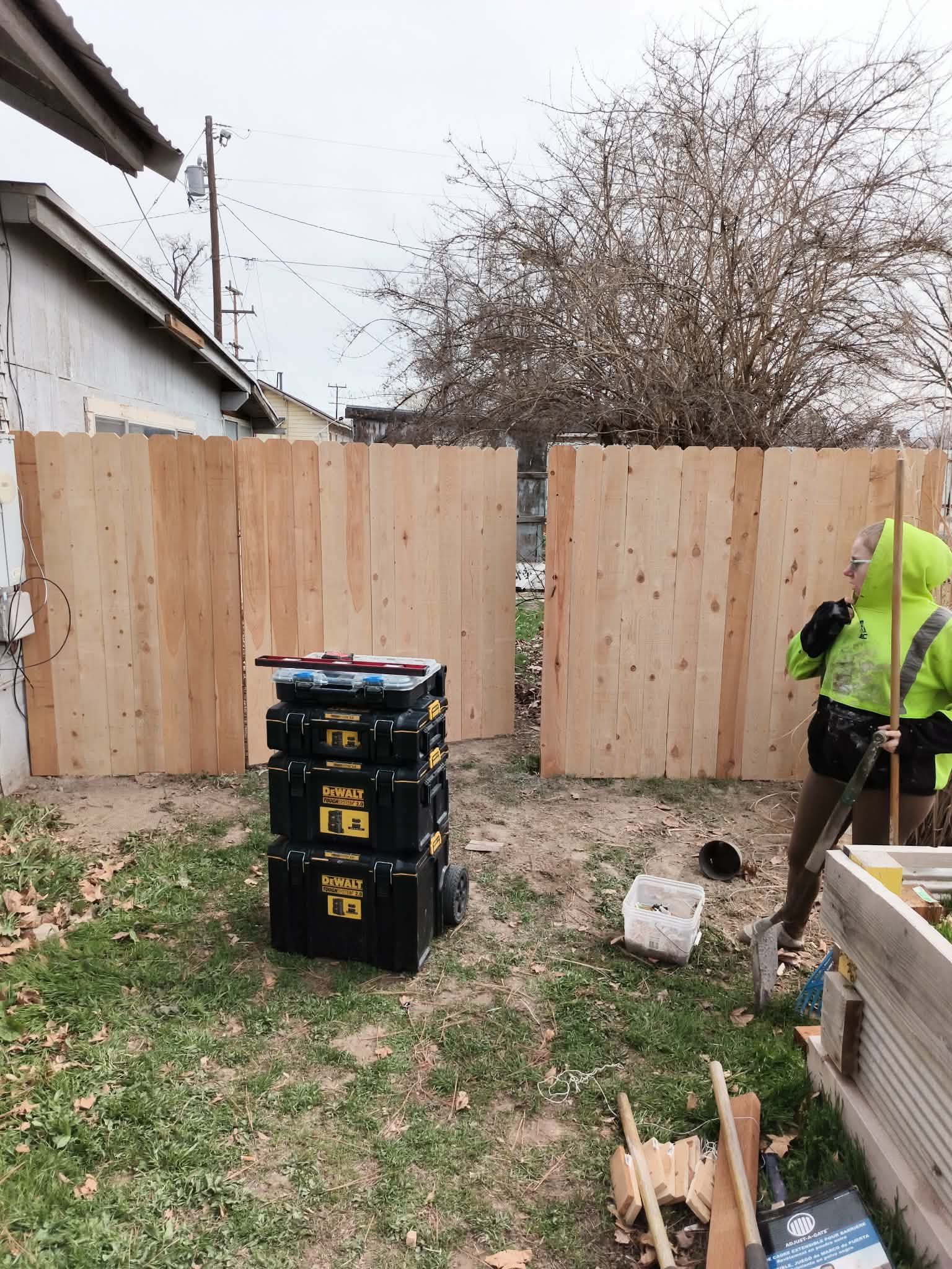 Wooden fence panels being installed in a backyard; worker in a neon jacket stands at right.