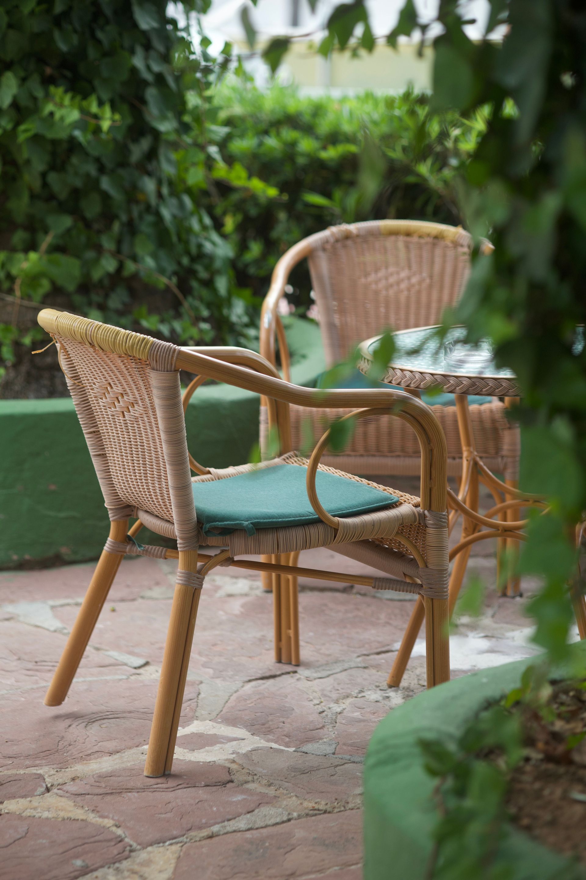 Two wicker chairs with teal cushions on a stone patio, surrounded by lush green plants.