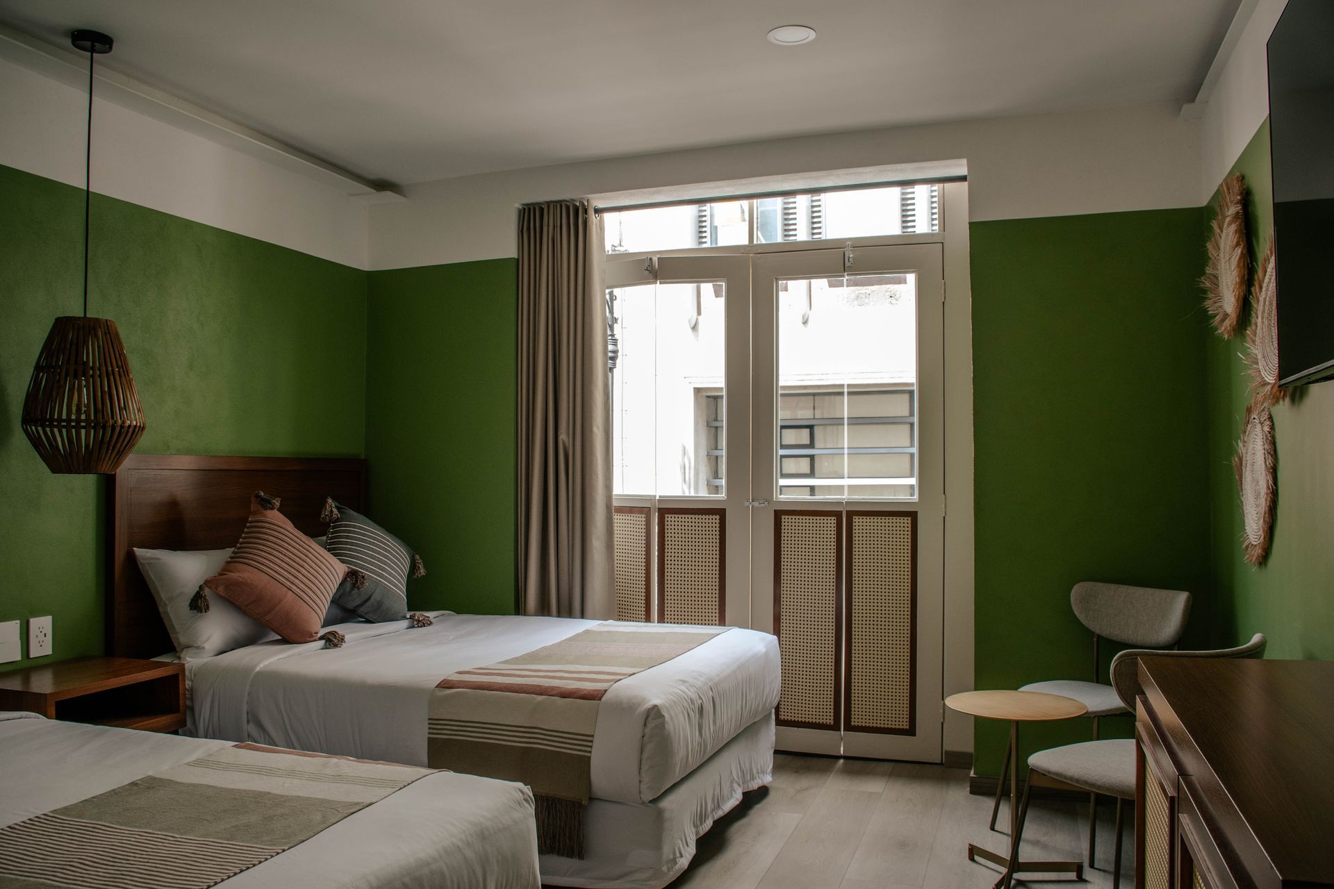 Dormitorio con paredes verdes, dos camas, una puerta acristalada que da al balcón y una mesita con una silla.
