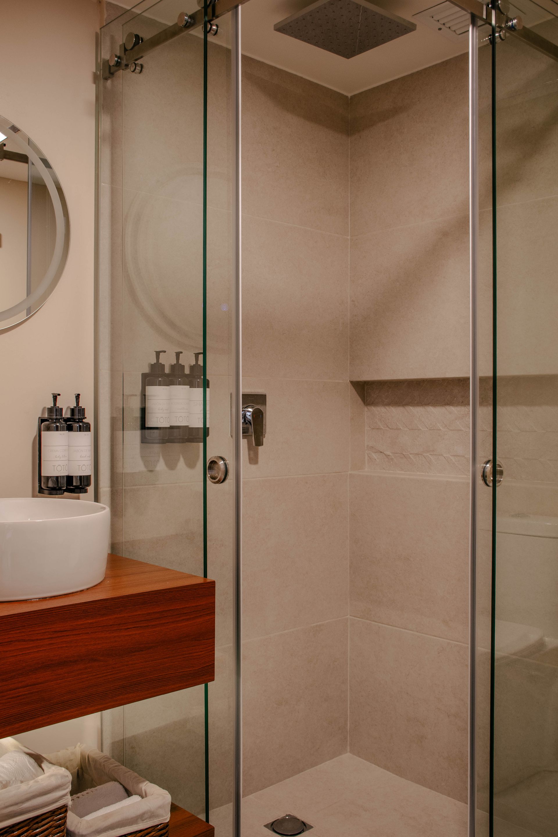Cabina de ducha de cristal con paredes de azulejos beige, tocador de madera y artículos de aseo en un baño