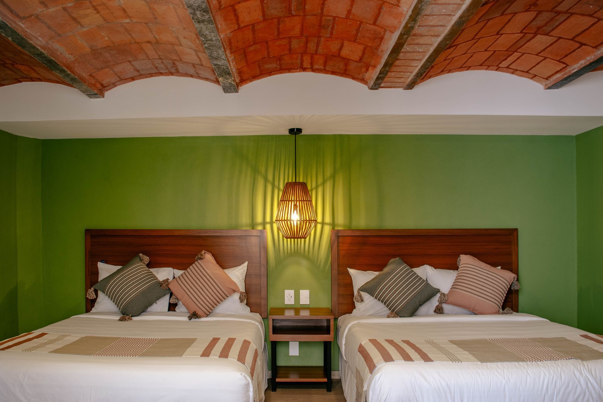 Dos camas individuales en una habitación verde con techo abovedado de ladrillo y una lámpara colgante entre ellas