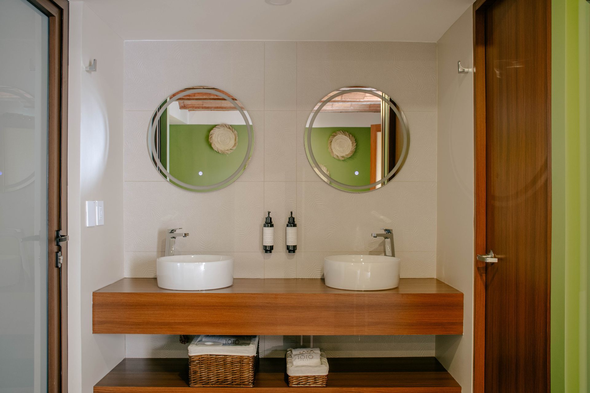 Tocador de baño moderno con dos espejos redondos, lavabos sobre encimera blancos y detalles de madera