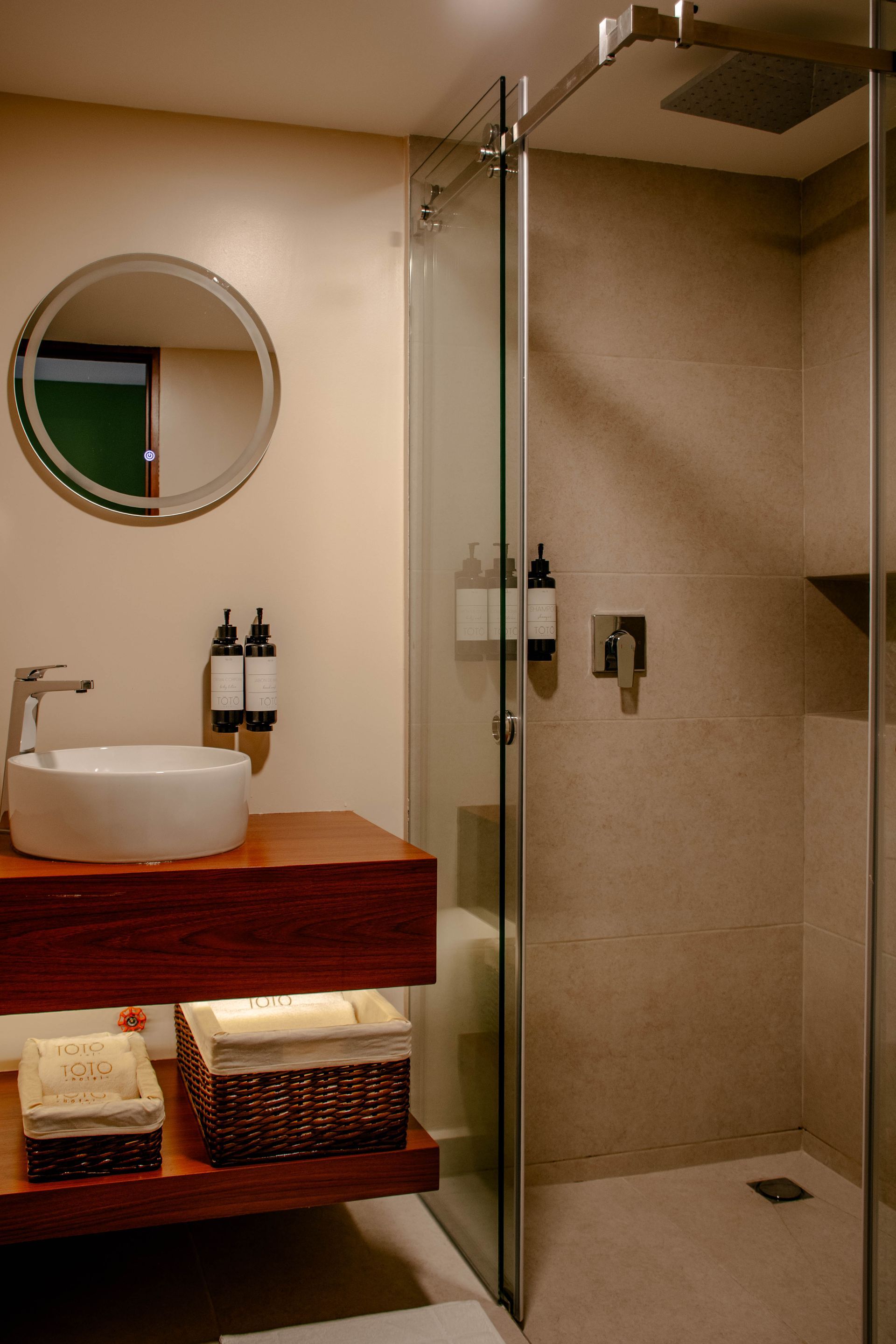 Baño moderno con ducha de cristal, espejo redondo, mueble de madera y cestas de mimbre