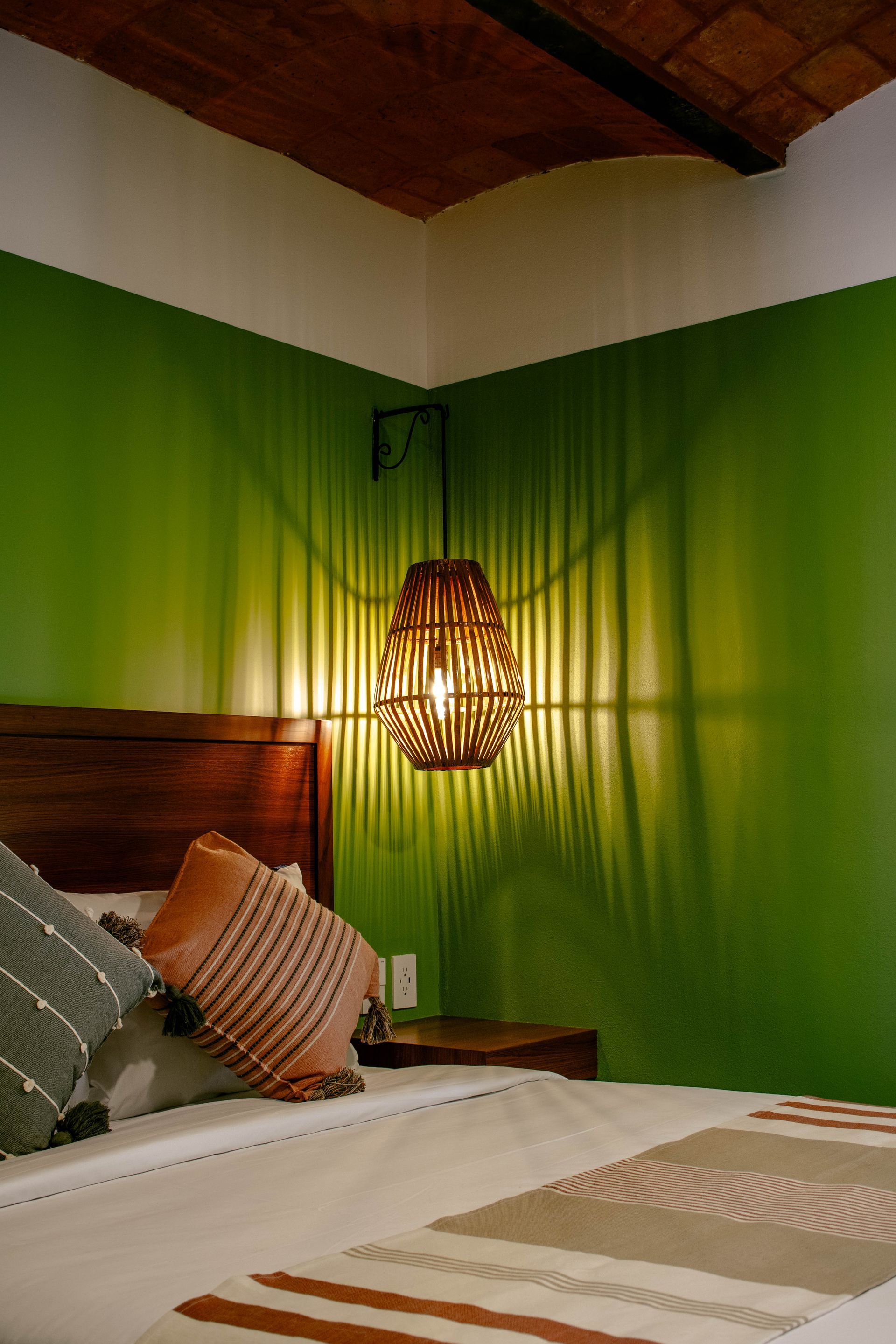Acogedor rincón de dormitorio con paredes verdes, cabecero de madera, almohadas a rayas y una lámpara colgante de mimbre