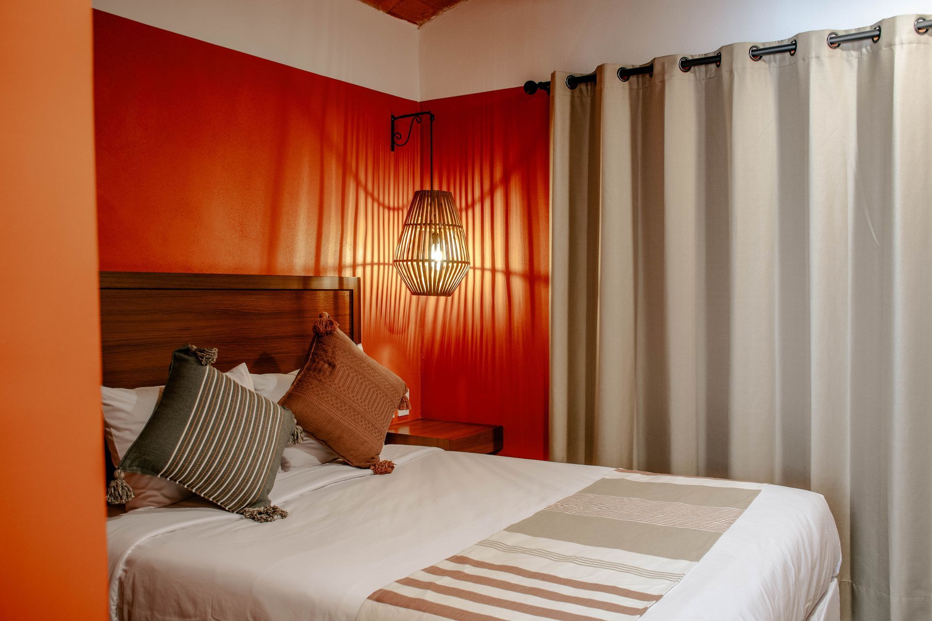Acogedor dormitorio con una pared de acento roja, una cama con almohadas y cortinas beige junto a una lámpara colgante
