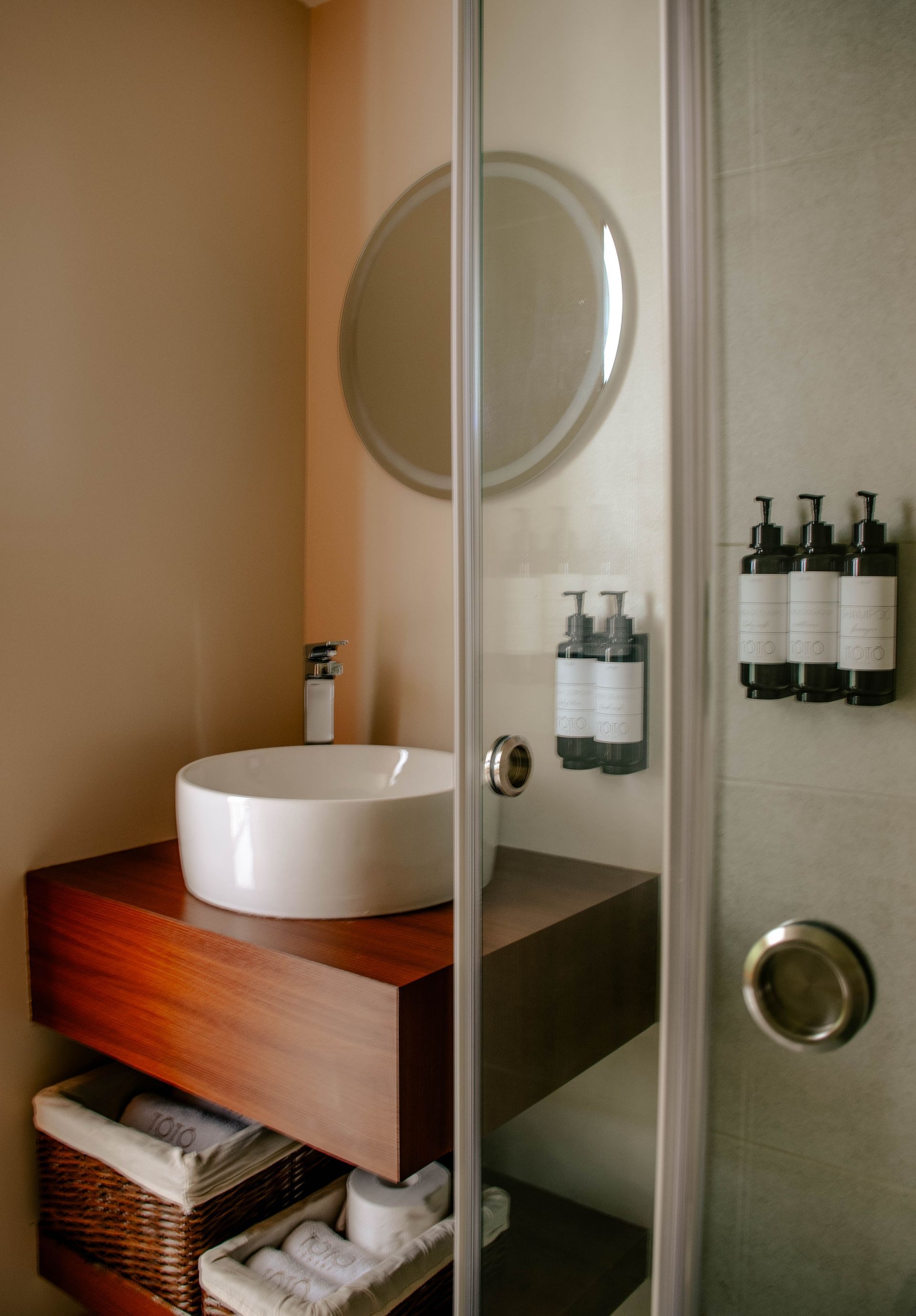 Baño moderno con ducha de cristal, espejo redondo y lavabo blanco sobre un mueble de madera
