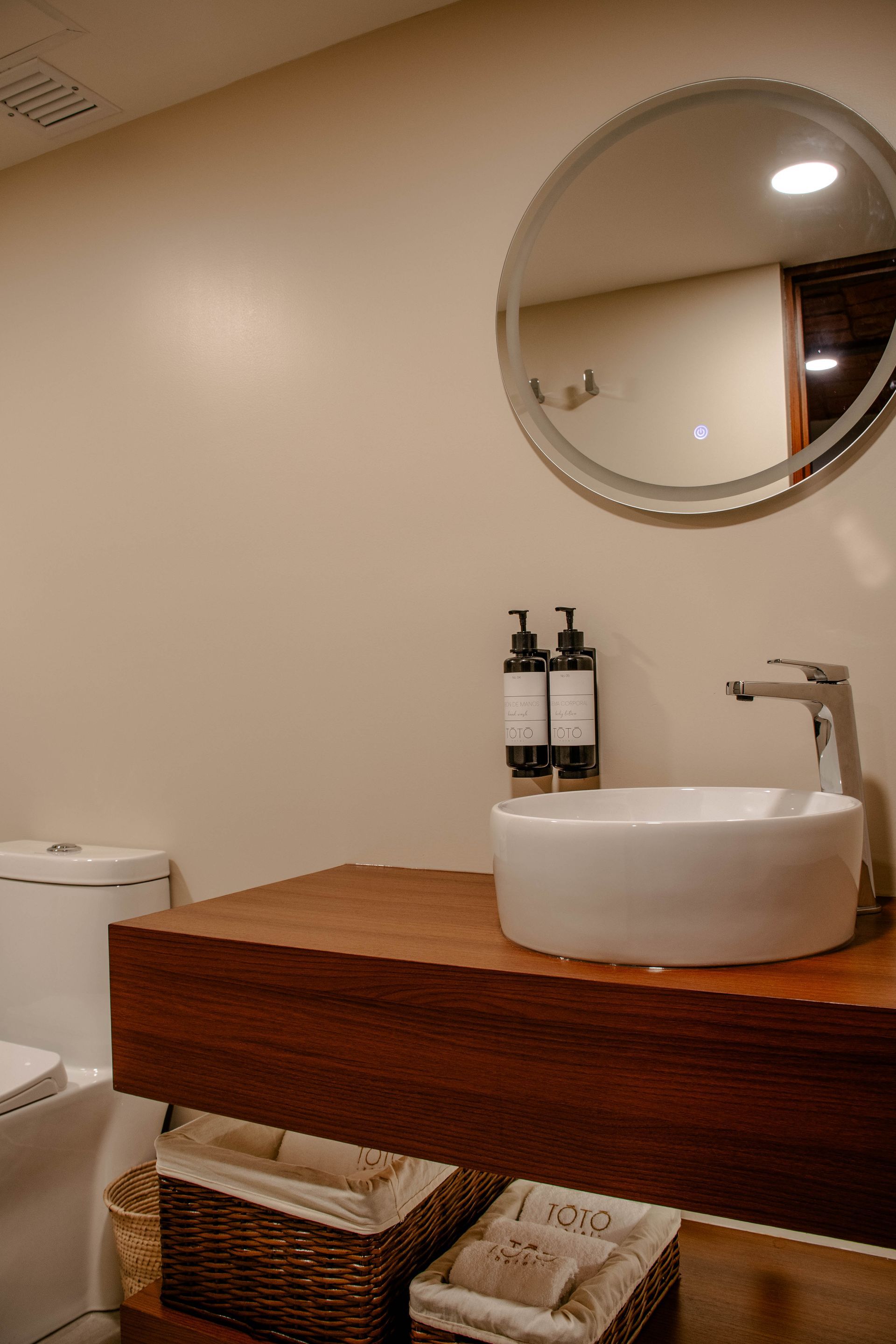 Baño moderno con espejo redondo, lavabo sobre encimera blanco, mueble de madera y cestas de mimbre para guardar cosas.