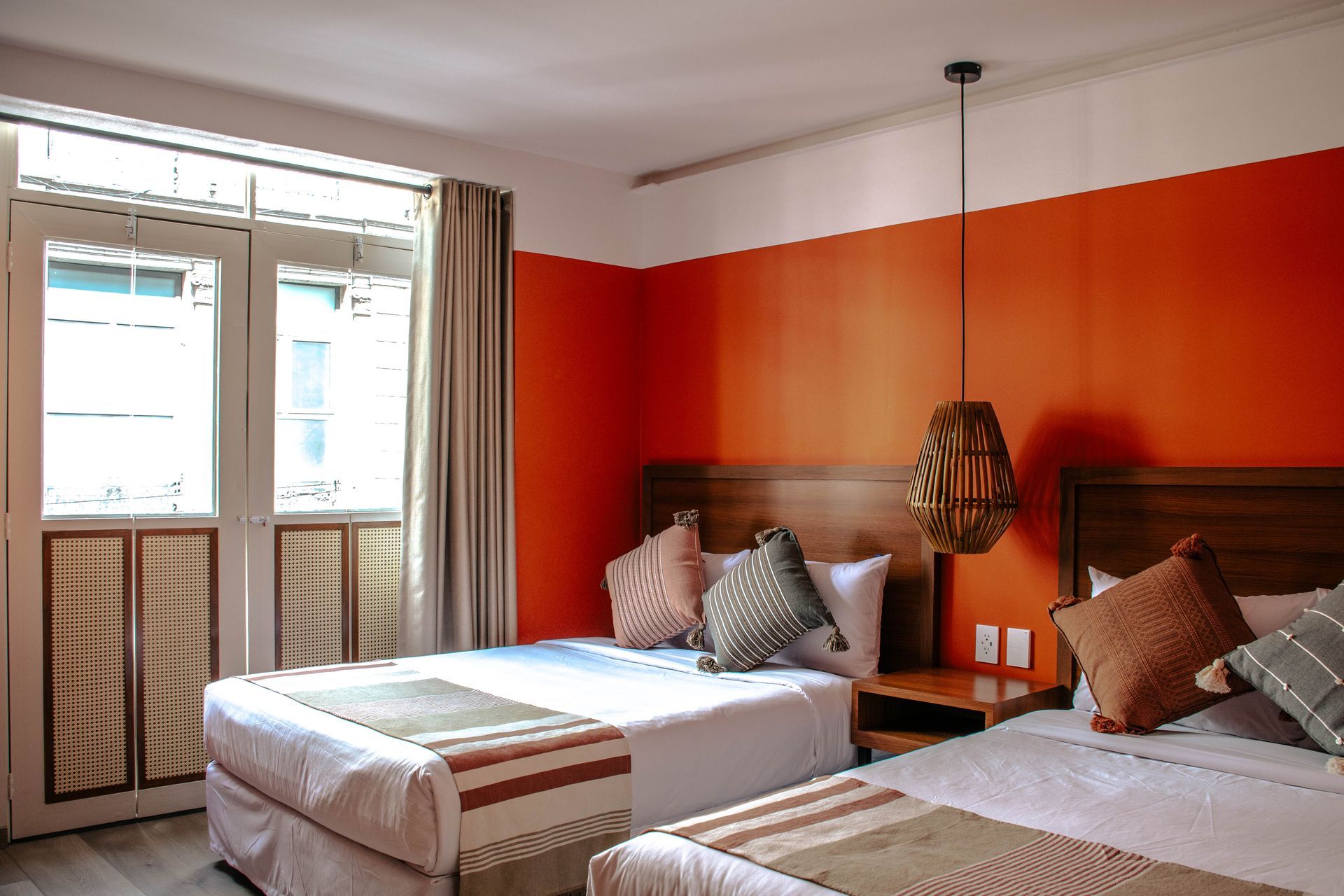 Dormitorio luminoso con dos camas, paredes naranjas y una ventana con cortinas y contraventanas