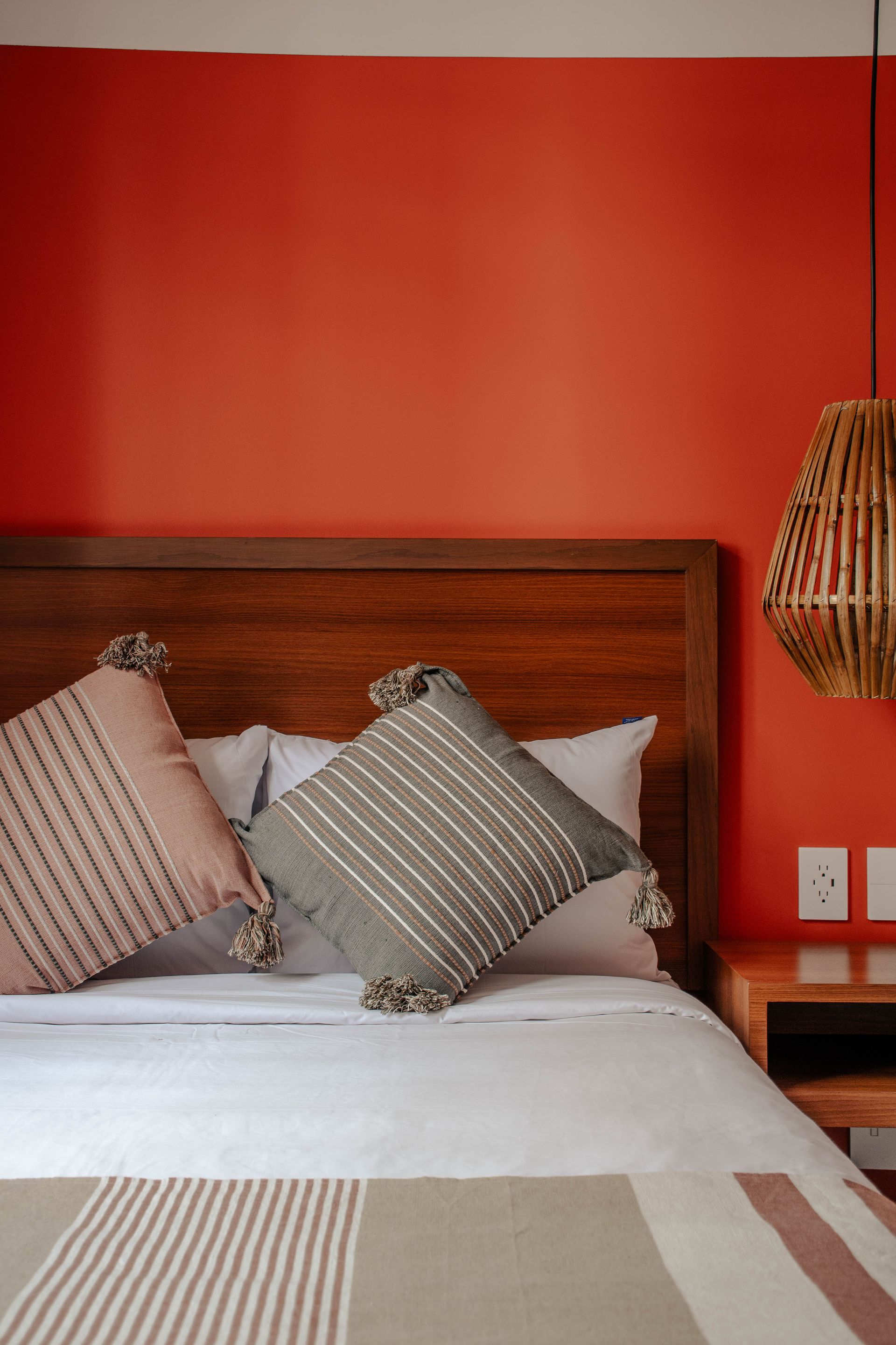 Cama con almohadas a rayas en una habitación con una pared color coral y una lámpara colgante de mimbre