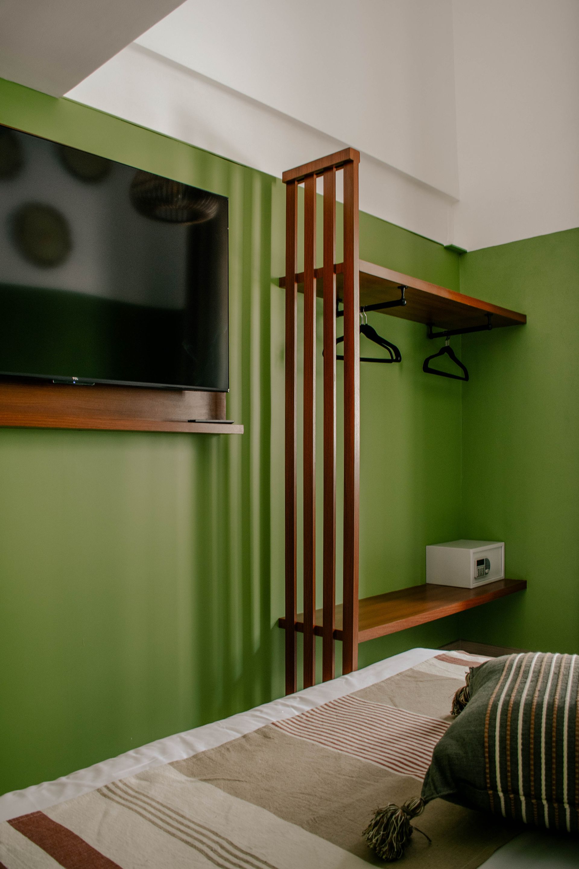 Dormitorio pequeño y verde con una cama, un televisor montado en la pared, un perchero de madera y estanterías.