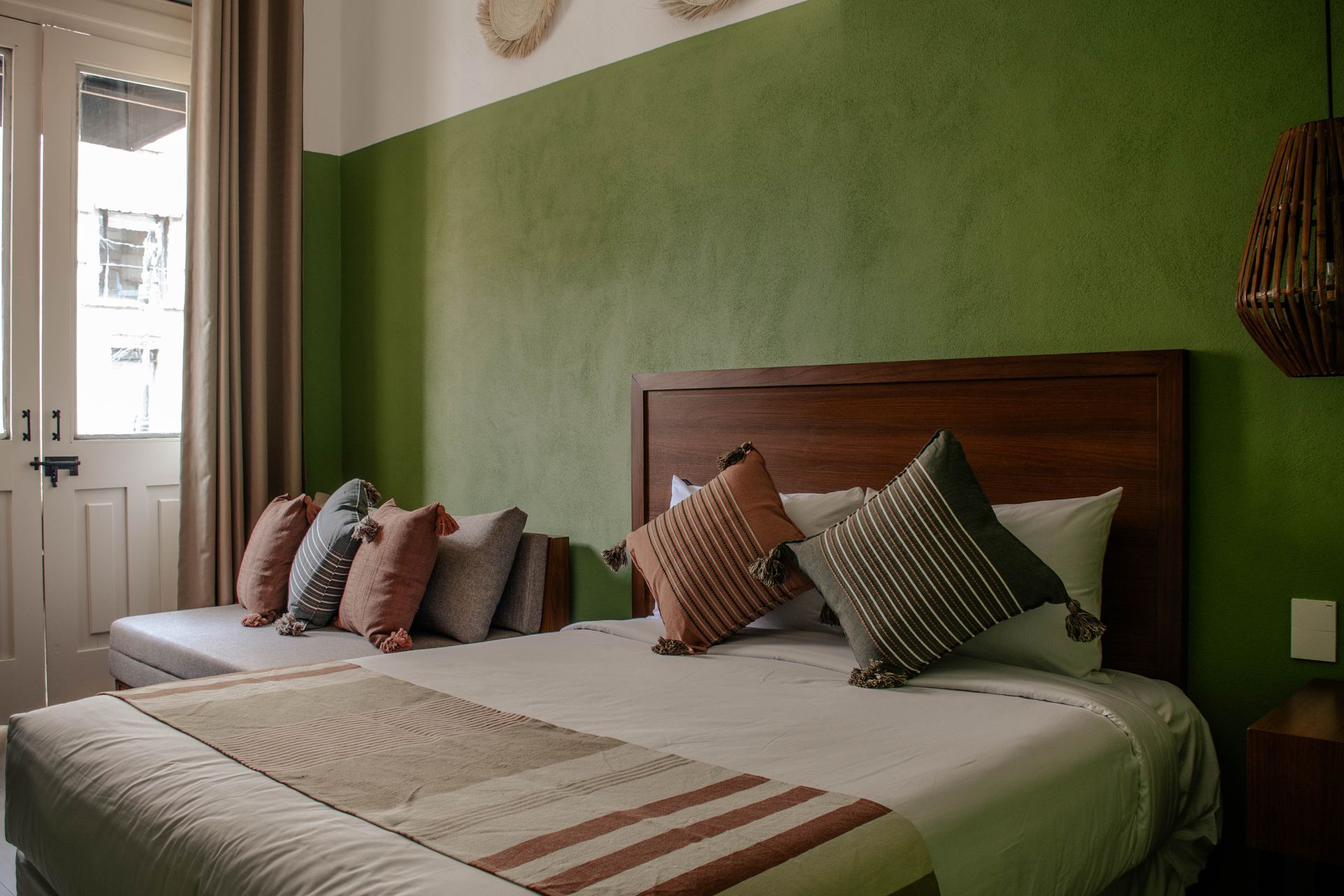Una cama bien hecha con almohadas estampadas en un dormitorio verde, junto a una ventana y una puerta blanca.