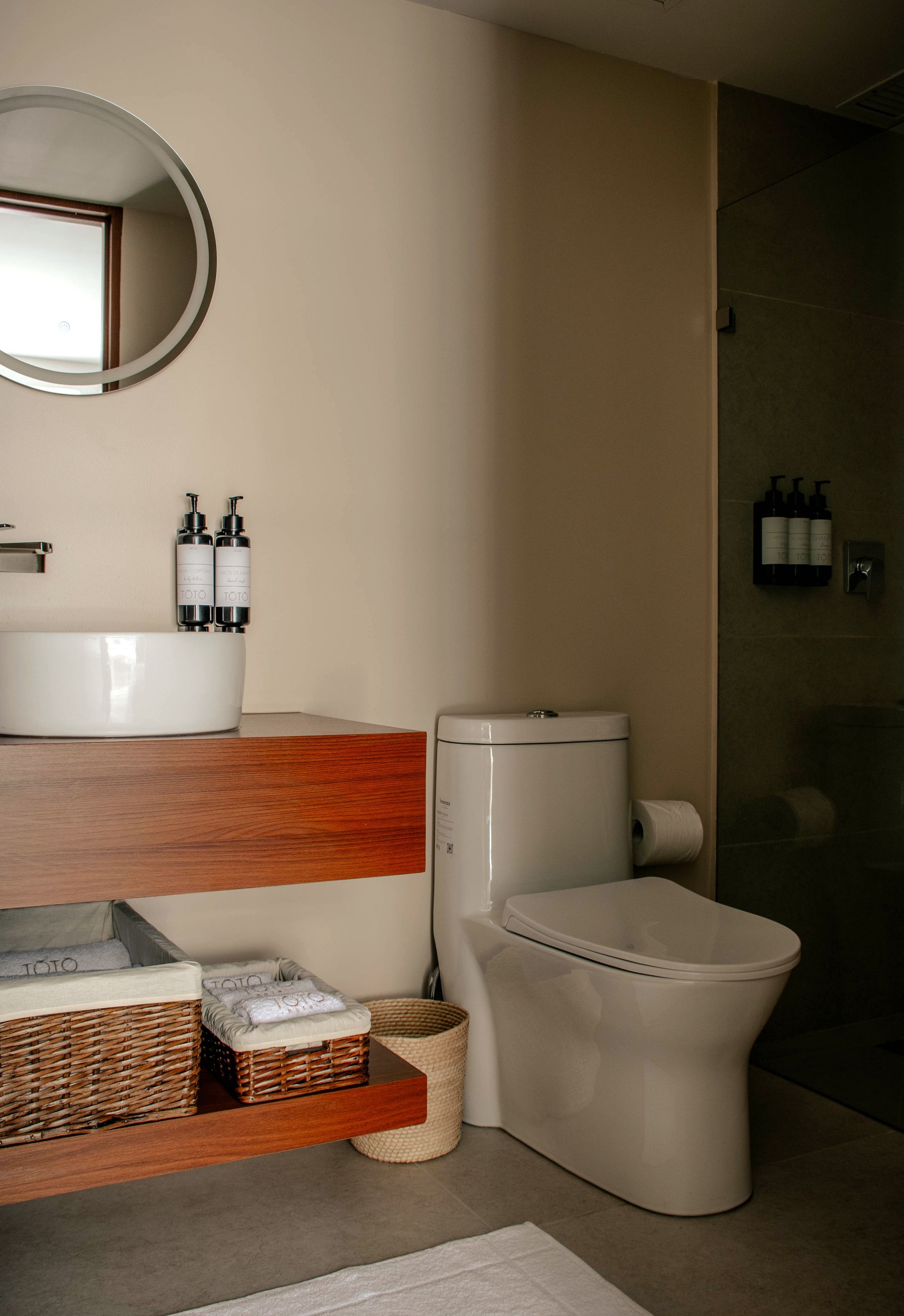Baño moderno con mueble de madera, espejo redondo, lavabo blanco, inodoro y cestas de mimbre