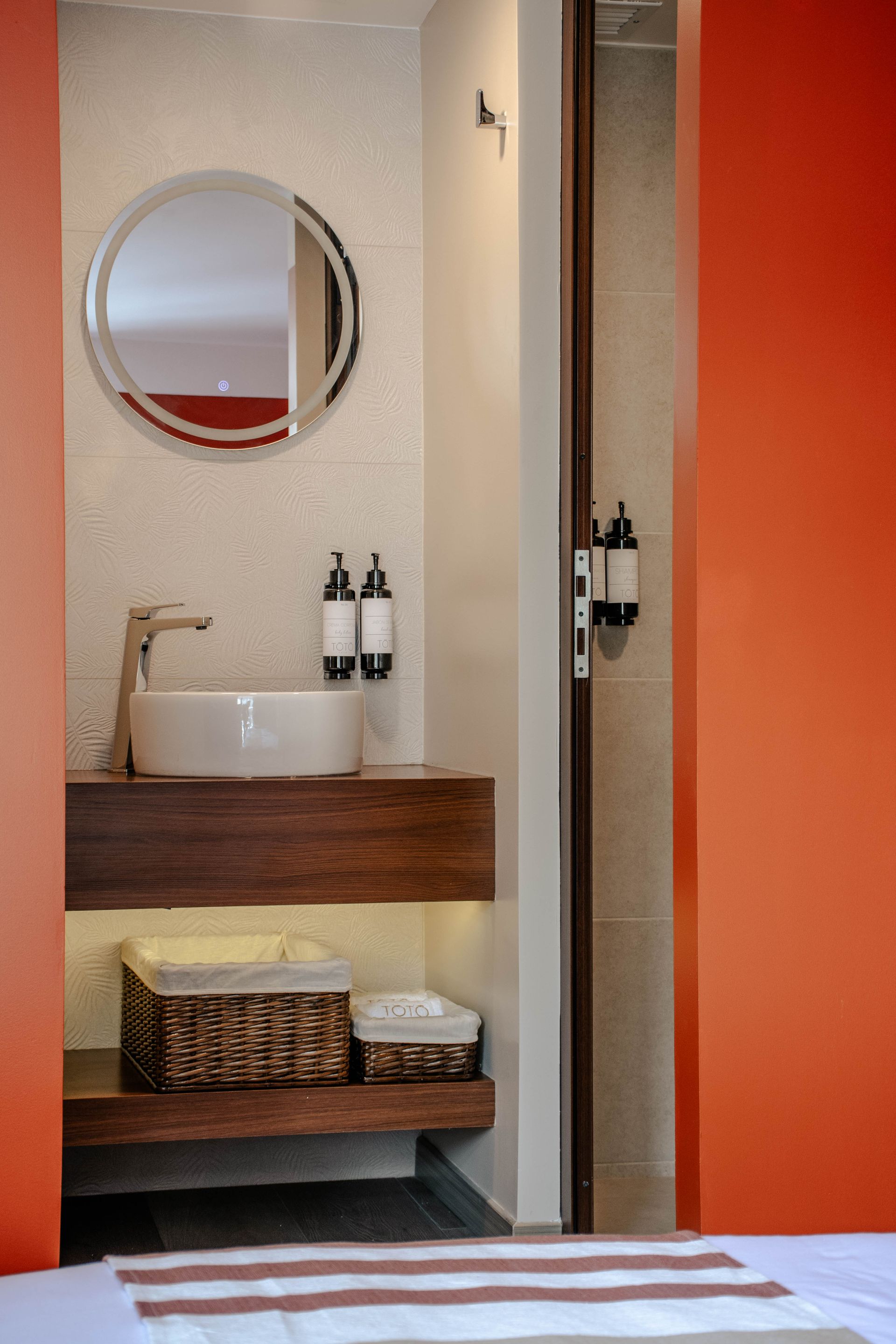 Baño moderno con paredes naranjas, espejo redondo, lavabo, ducha y estante con cestas de mimbre.