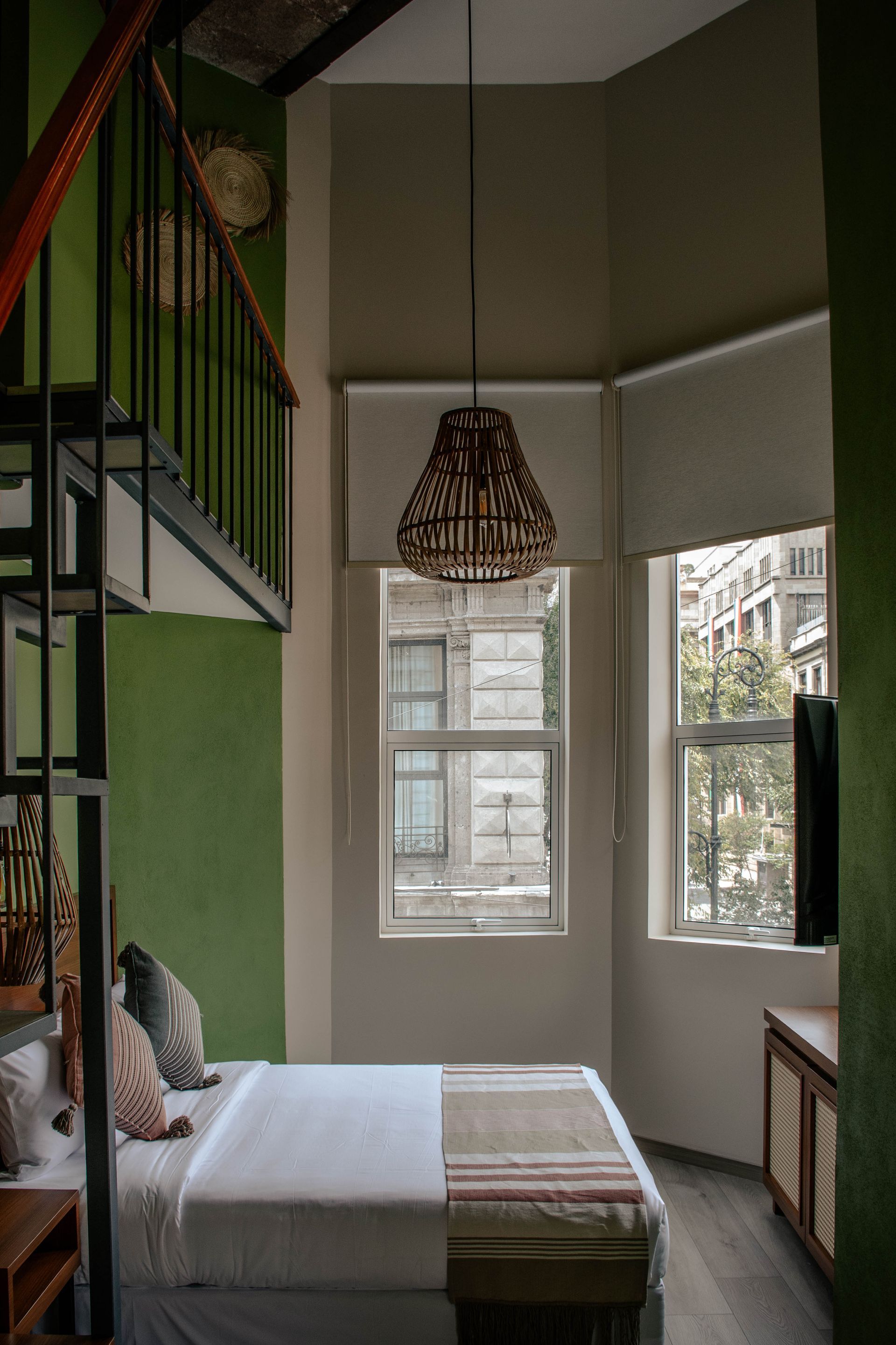Dormitorio estrecho con paredes verdes, una cama, una escalera, una lámpara colgante y dos ventanas con luz natural.