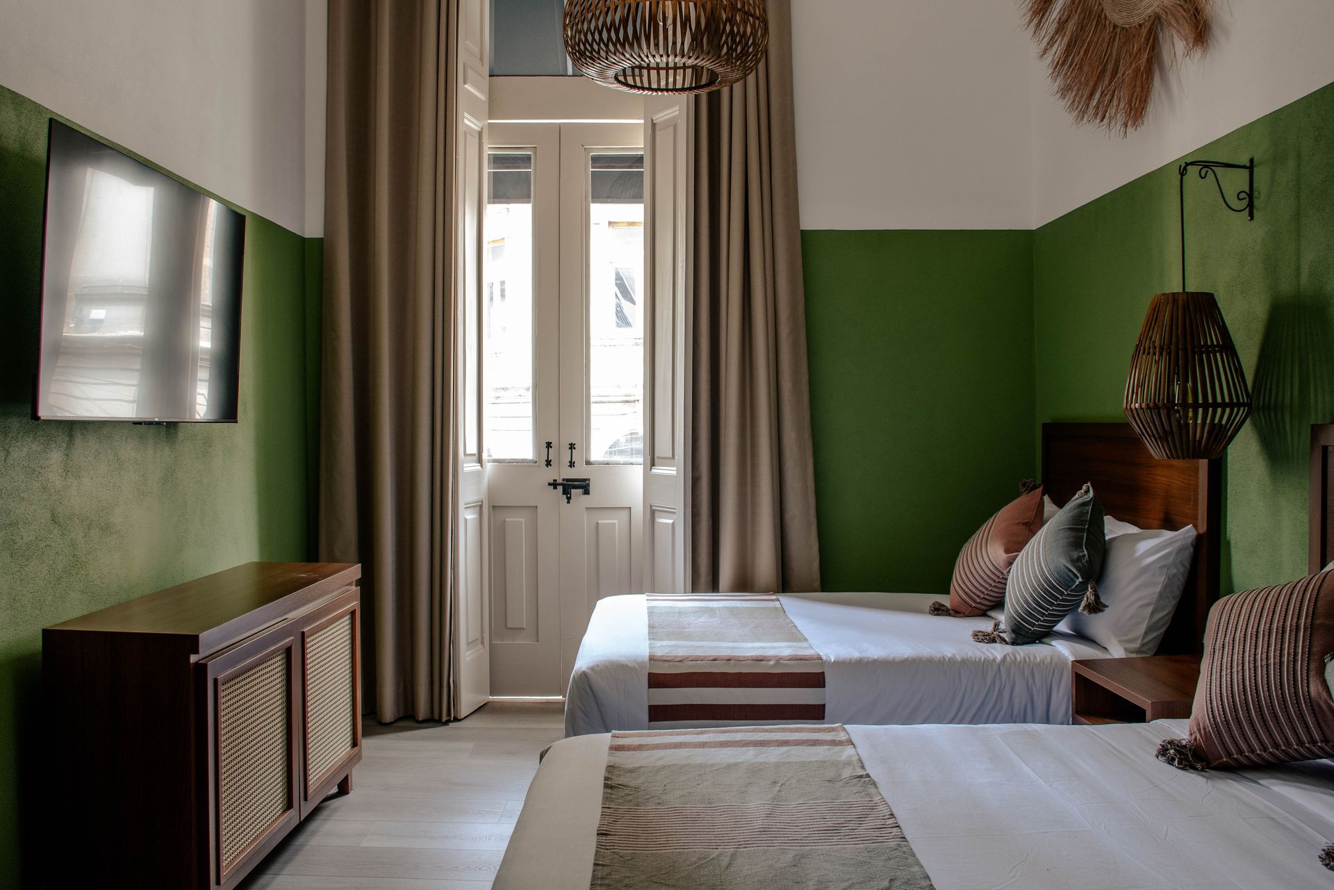 Dormitorio pequeño con dos camas individuales, paredes verdes, una puerta con ventana y un televisor montado en la pared.