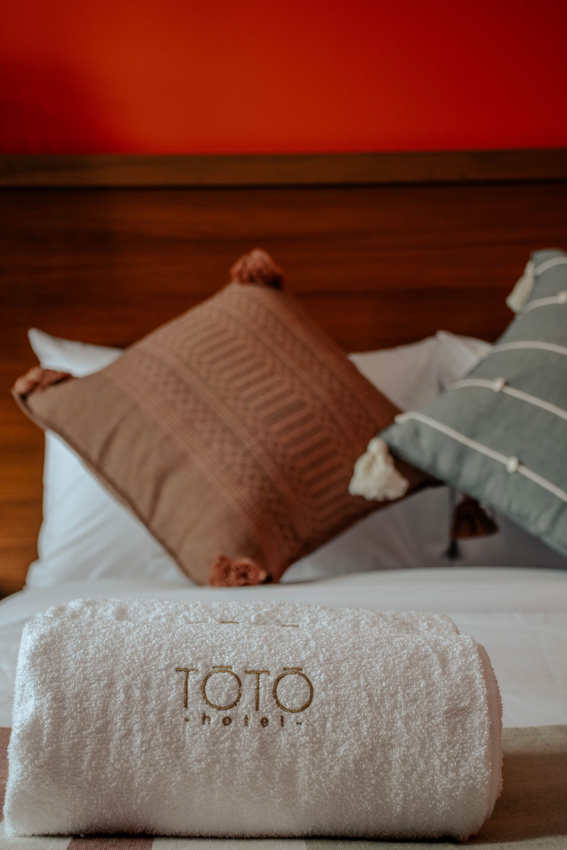 Cama con cabecero de madera, cojines en tonos óxido y gris, y una toalla blanca enrollada delante