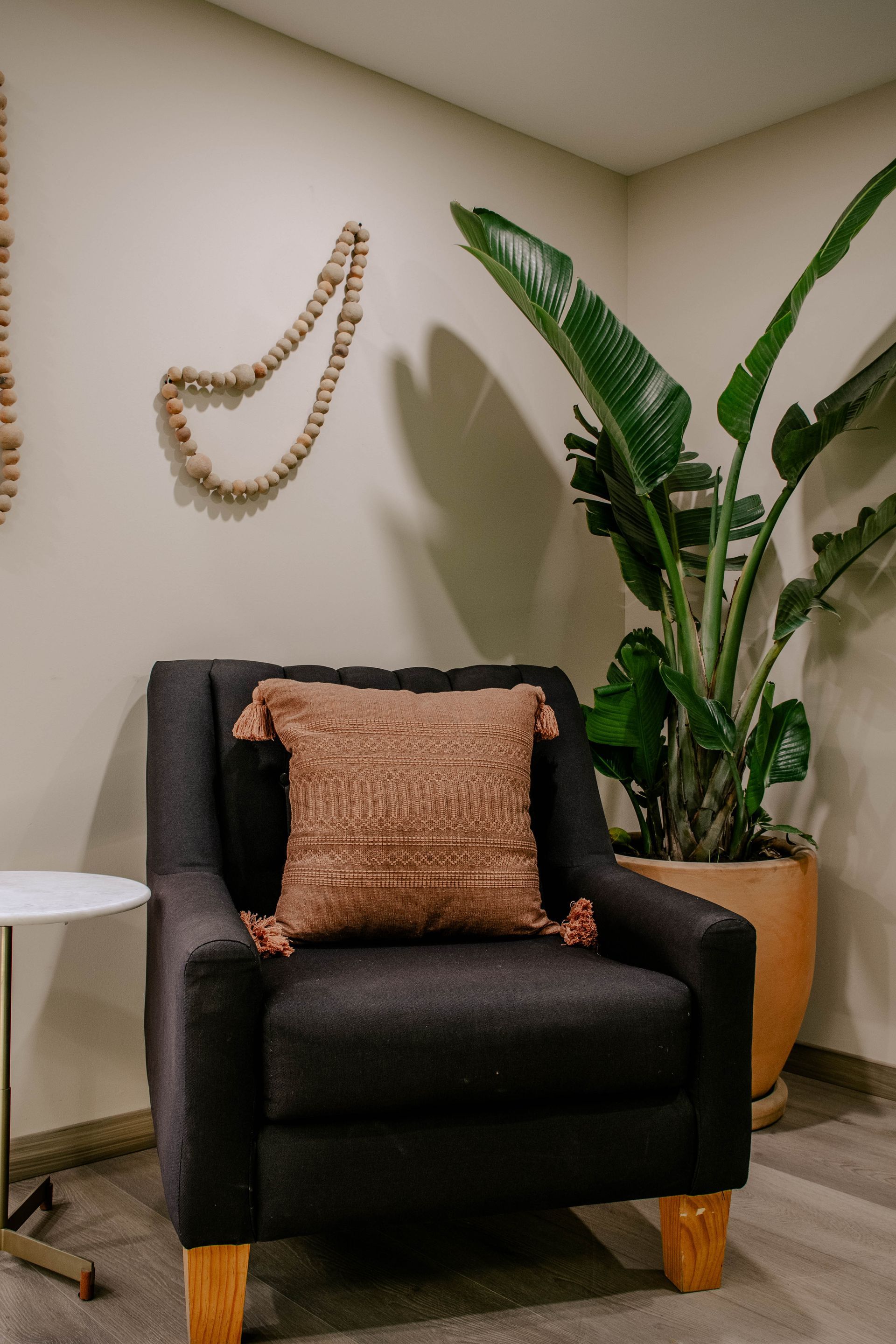 Rincón acogedor con sillón negro, cojín rosa, planta alta en maceta y decoración de pared en una habitación beige