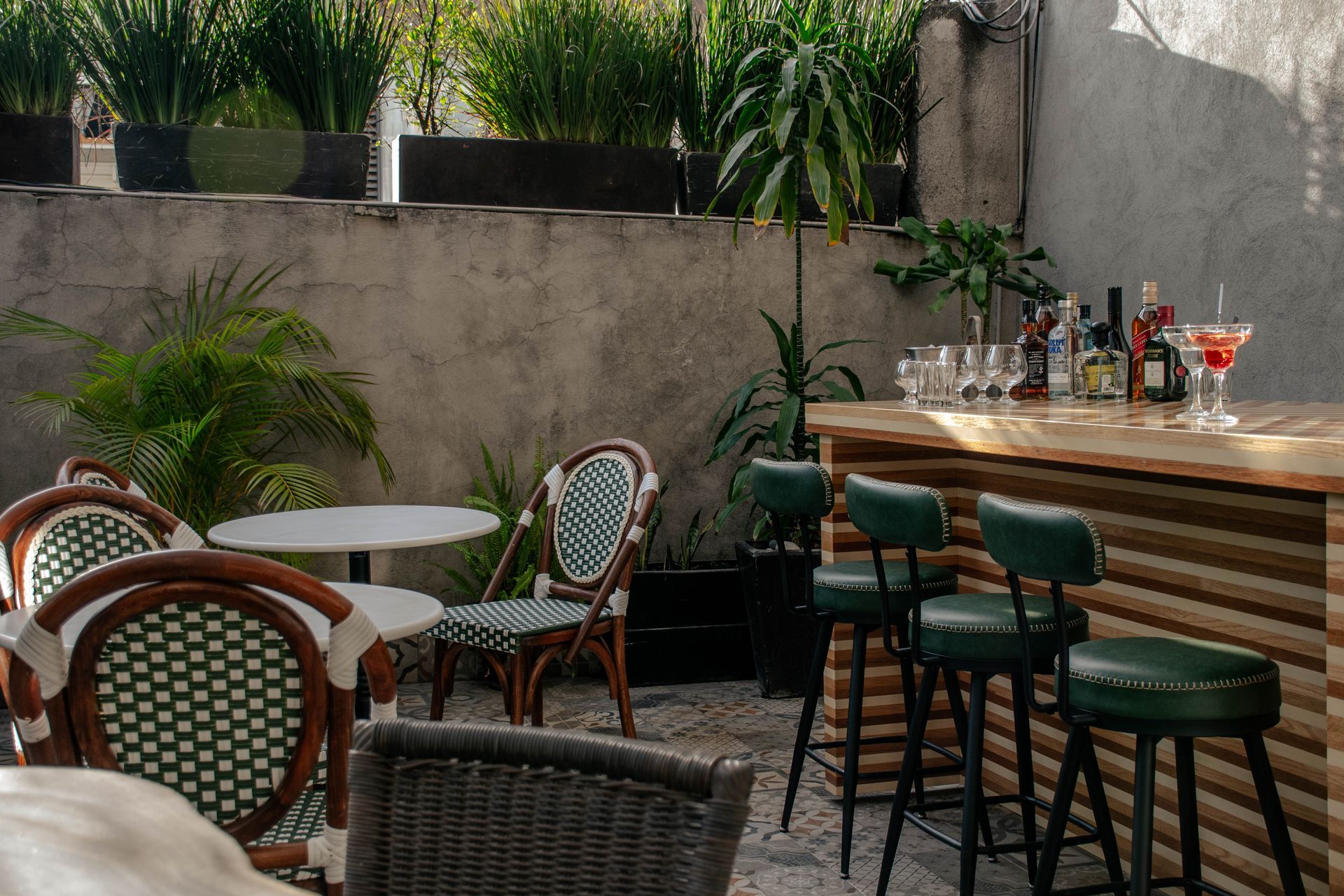 Bar al aire libre en un patio con sillas de mimbre, mesitas, plantas en macetas y una barra de madera con taburetes