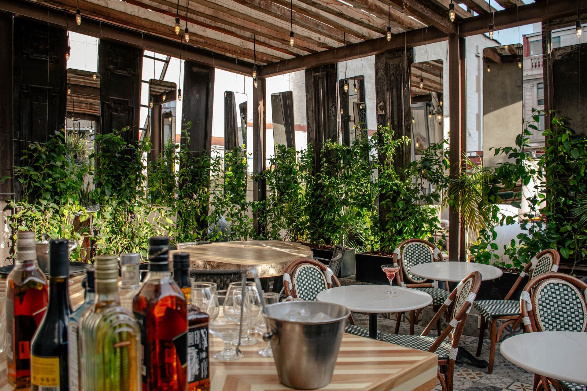Patio al aire libre con techo de listones de madera, mesa de comedor, plantas y una pequeña barra sobre un suelo de baldosas.