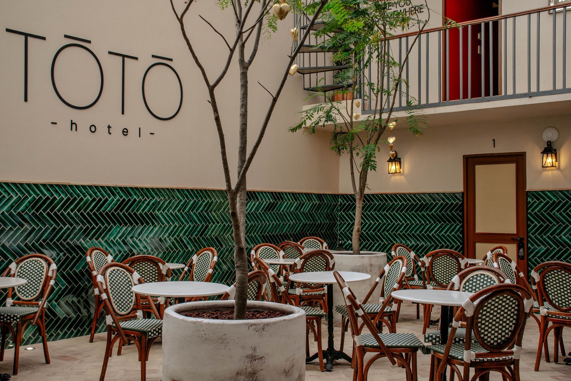 Patio exterior de hotel con mesas de cafetería, sillas de mimbre, un árbol en una maceta y un letrero de pared del hotel TOTO
