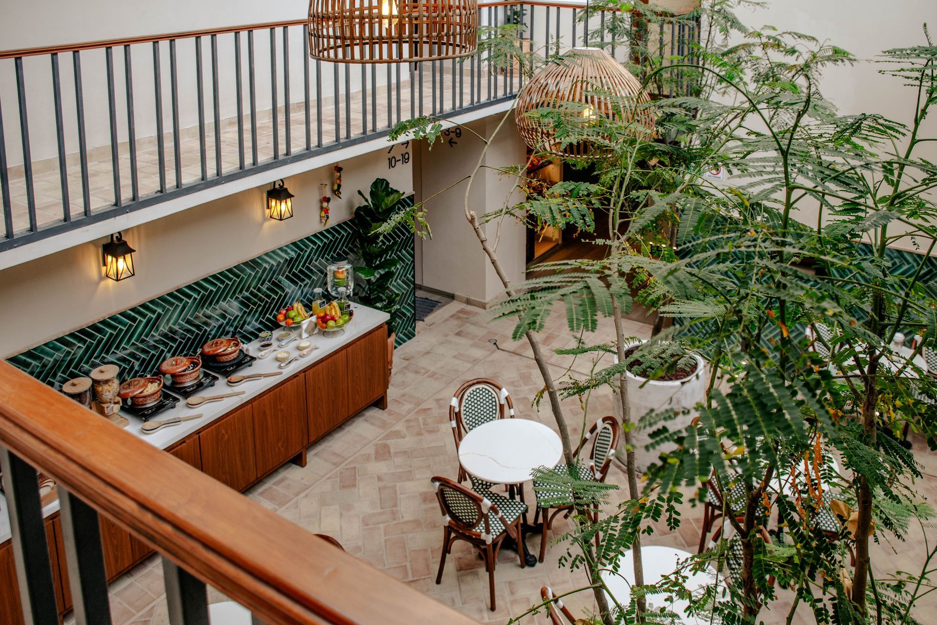 Luminoso patio interior con una mesa de bufé de madera, una mesa redonda de cafetería y exuberantes plantas bajo un balcón.
