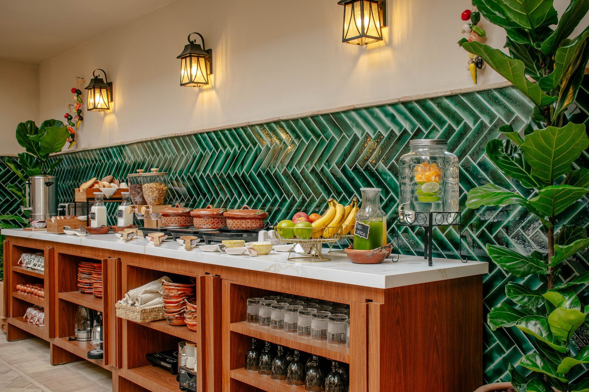 Mesa de salón repleta de platos variados para el desayuno, pasteles, fruta y bebidas, junto a un sofá y una lámpara.