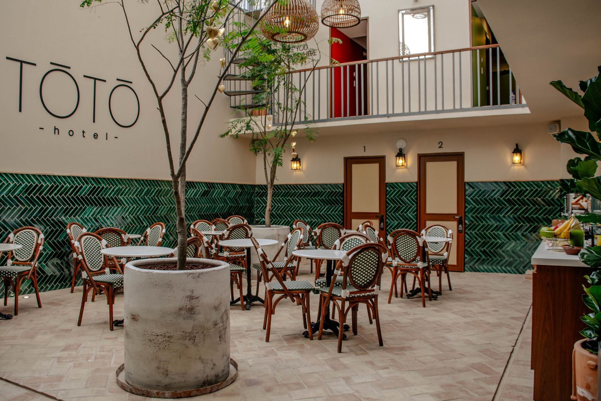 Terraza de una cafetería al aire libre con mesas, sillas de mimbre, plantas y un balcón arriba, con el rótulo «TOTO»