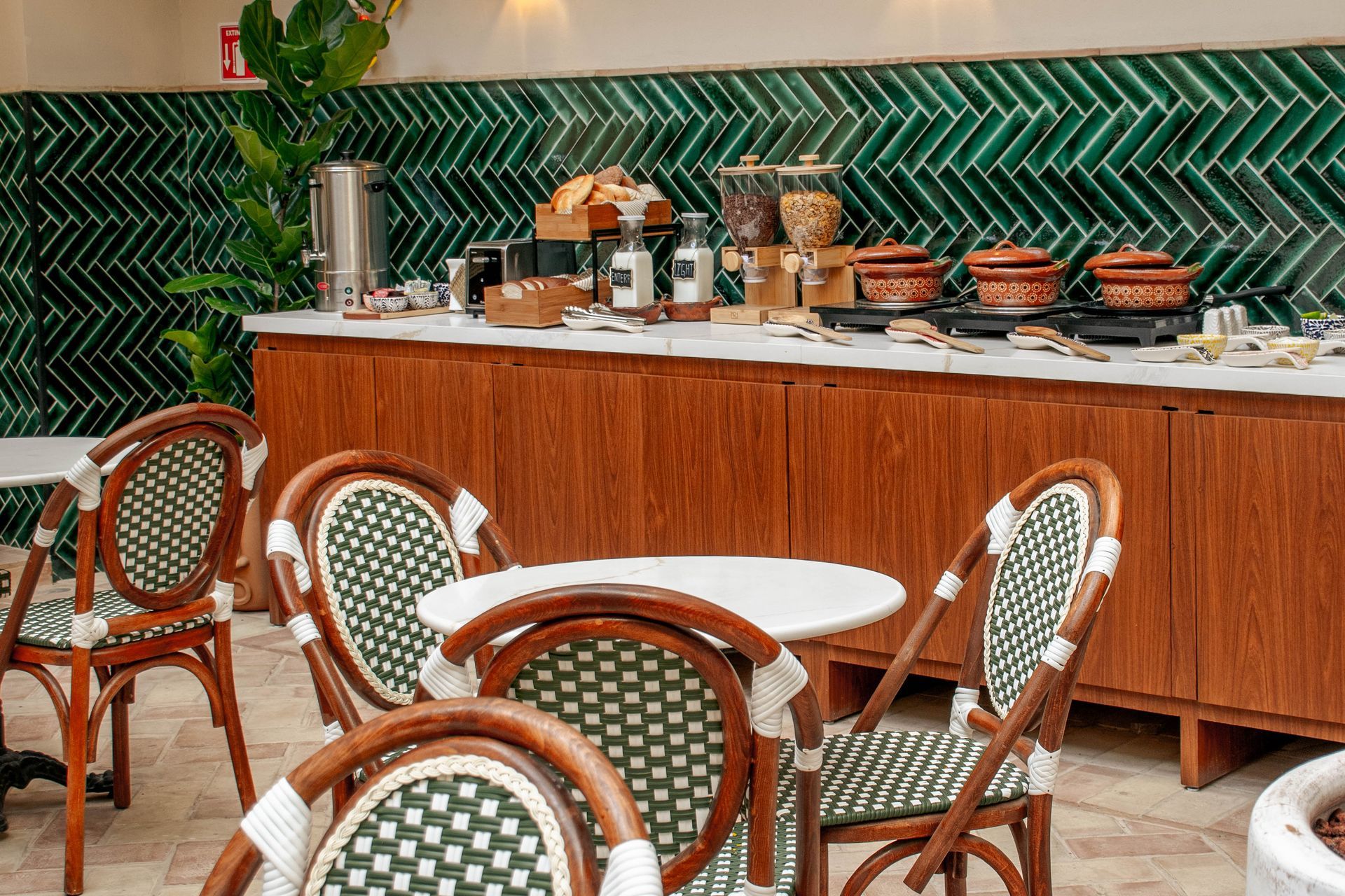 Mesas de terraza de un café, con sillas de mimbre y mesas blancas frente a un mostrador de madera tipo bufé