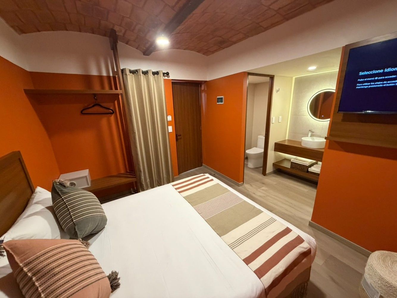 Habitación de hotel moderna con cama, paredes naranjas, techo de madera, armario y vista al área del baño.