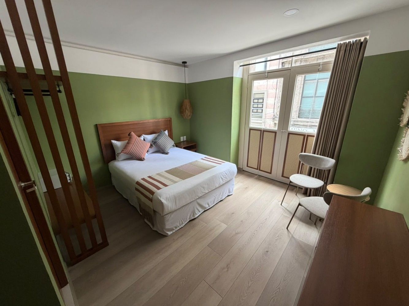 Habitación luminosa con paredes verdes, suelo de madera, cama, puertas con ventana y una silla pequeña.