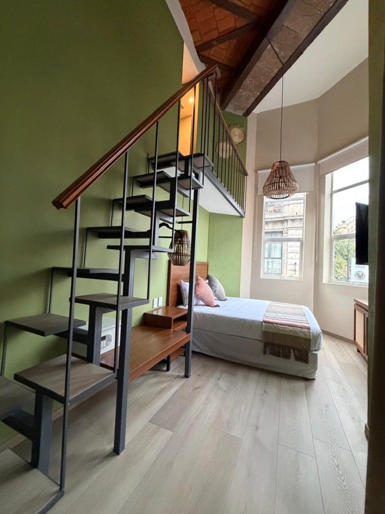 Habitación tipo loft con escalera negra, pared verde, cama y gran ventanal con vistas a los árboles.