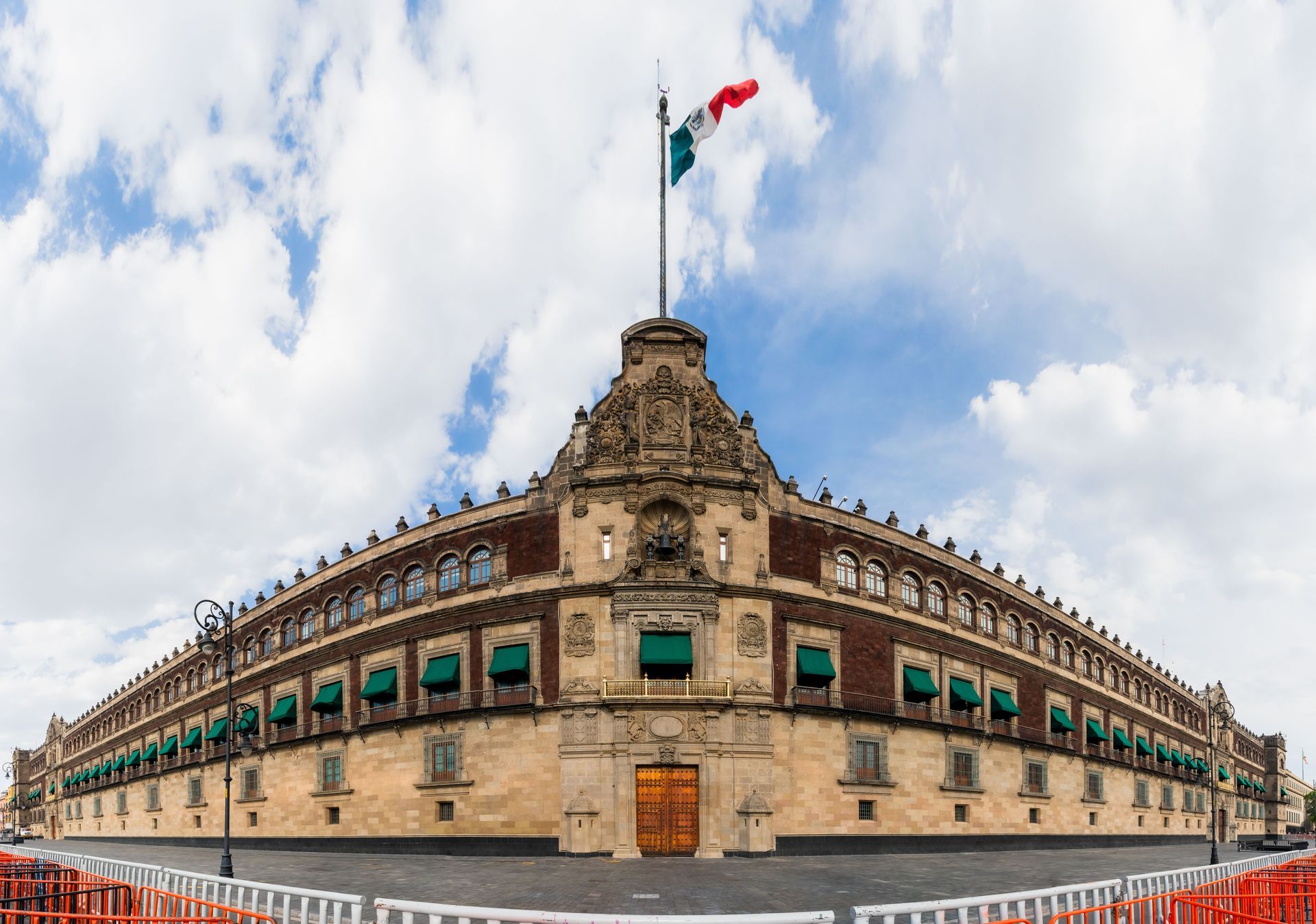 Edificio histórico de piedra con una bandera mexicana ondeando en lo alto, visto desde un ángulo bajo un cielo nublado.