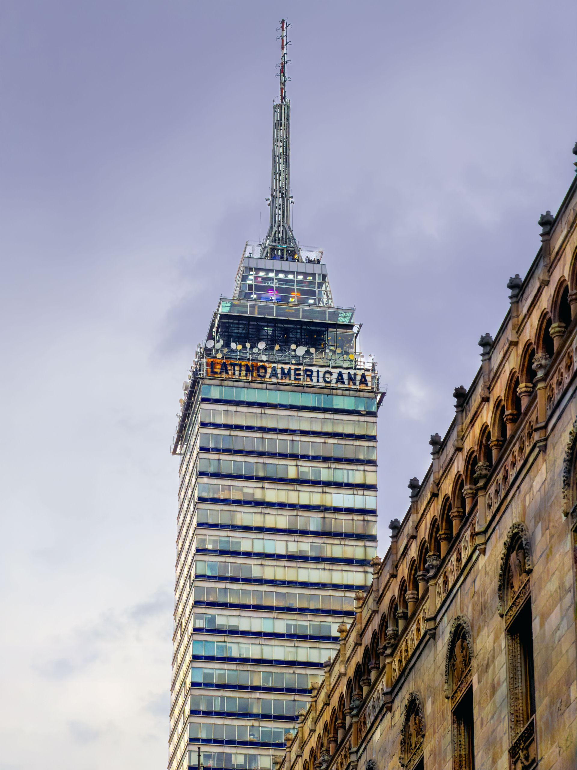 Un alto rascacielos moderno se recorta contra un cielo nublado, enmarcado por un edificio histórico ornamentado a la derecha.