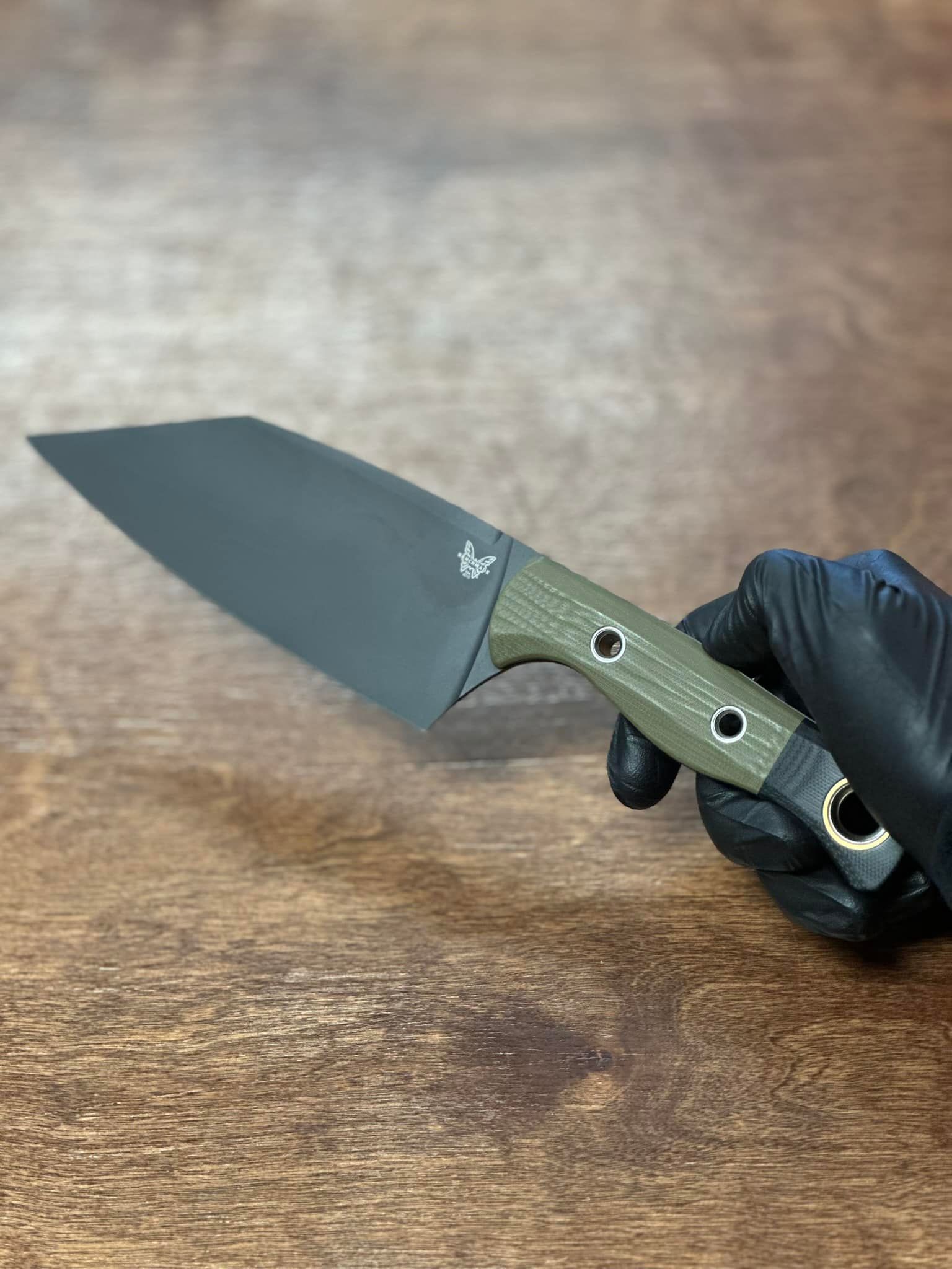 Black-gloved hand holding an olive-green folding knife on a wooden surface.